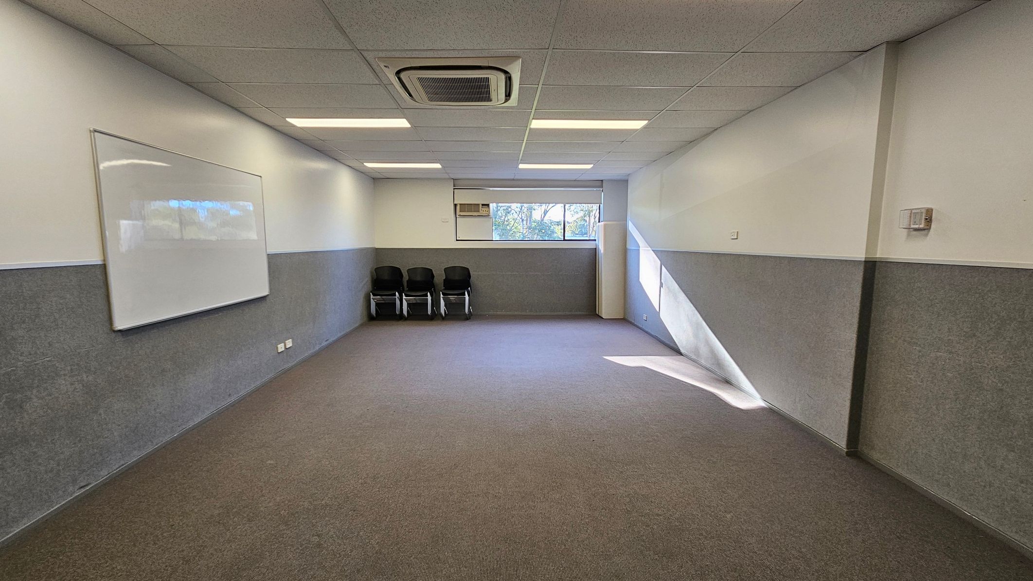 A rectangular room with a brown carpeted floor and a ceiling with recessed lighting and an air conditioning unit. On the left wall, there's a large whiteboard. At the far end, three stacked chairs are placed against the wall beneath a window, which lets in natural light. The walls are painted in two tones, with the lower half in grey. A rectangular room with a brown carpeted floor and a ceiling with recessed lighting and an air conditioning unit. On the left wall, there's a large whiteboard. At the far end, three stacked chairs are placed against the wall beneath a window, which lets in natural light. The walls are painted in two tones, with the lower half in grey.