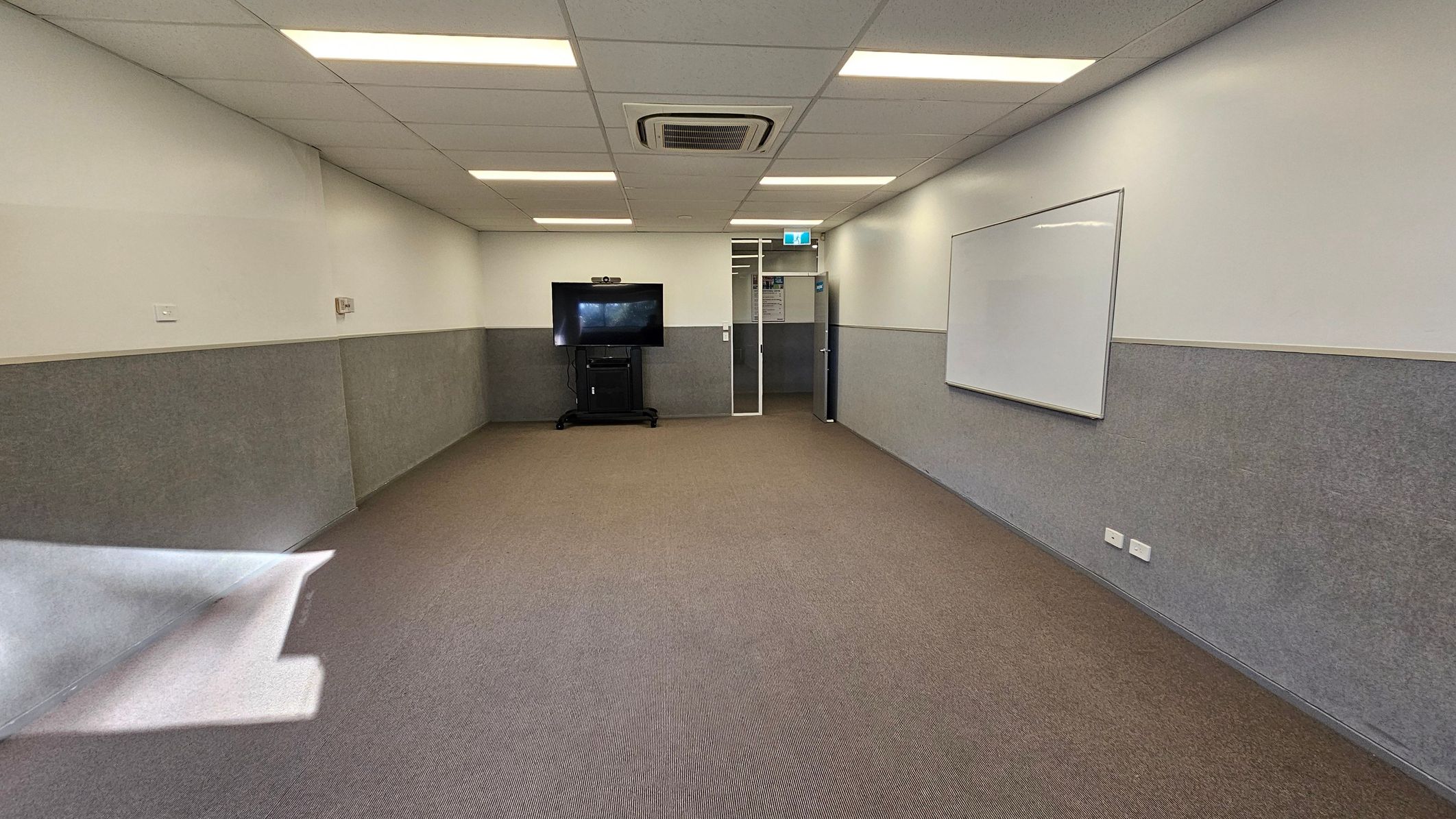 A rectangular room with a brown carpeted floor and a white ceiling featuring recessed lighting and an air conditioning unit. On the right wall, there's a large whiteboard. At the far end, a flat-screen TV is mounted on a stand. The walls are half-painted in grey and white. A door with a glass panel is located next to the TV, leading to another area. The room appears to be a meeting or classroom space. A rectangular room with a brown carpeted floor and a white ceiling featuring recessed lighting and an air conditioning unit. On the right wall, there's a large whiteboard. At the far end, a flat-screen TV is mounted on a stand. The walls are half-painted in grey and white. A door with a glass panel is located next to the TV, leading to another area. The room appears to be a meeting or classroom space.