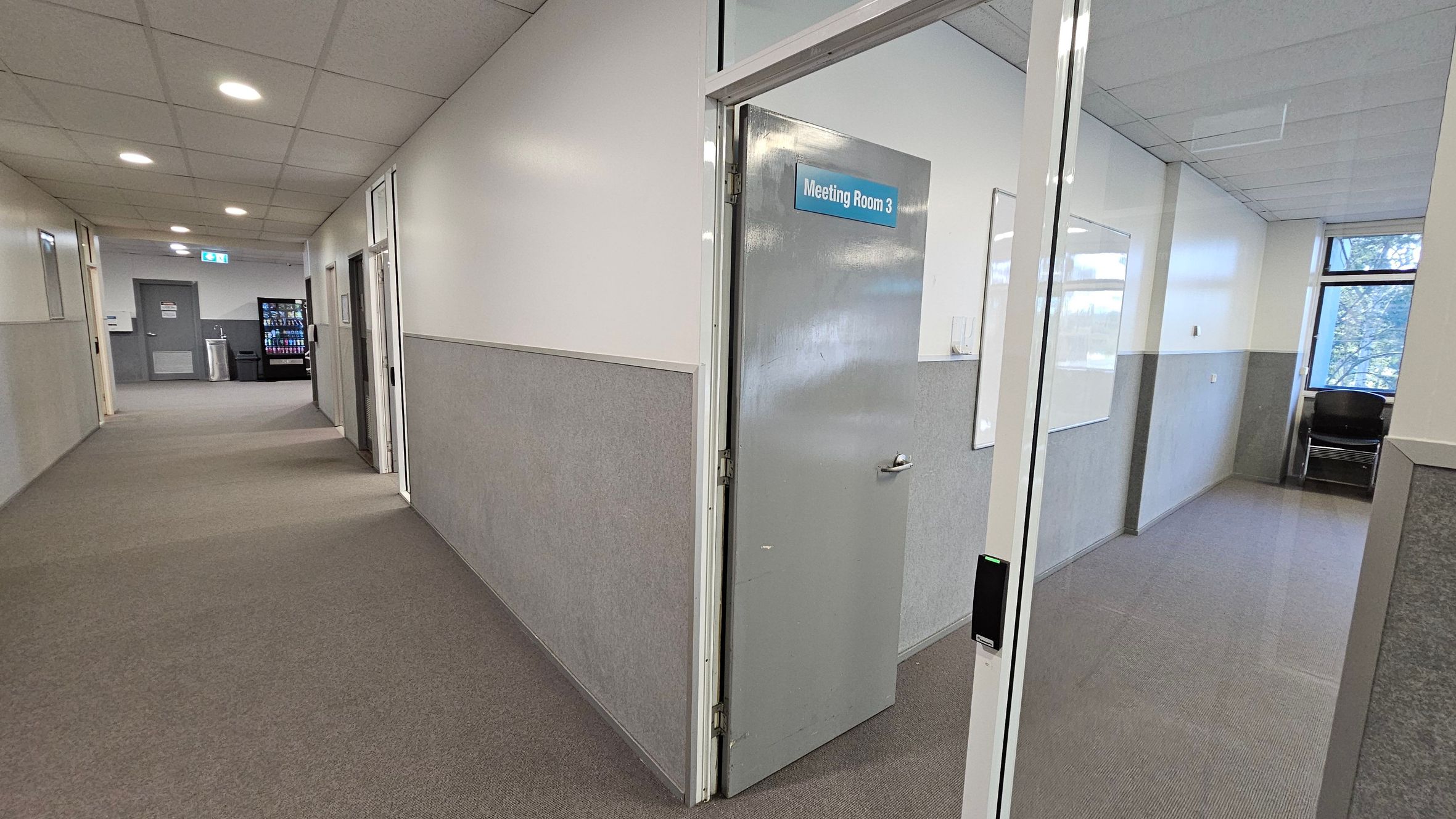 A corridor with beige carpet and white walls, featuring a grey door on the right labelled "Meeting Room 3." Inside the room, there's a whiteboard on the wall and a window with a view of trees. In the background, the corridor extends with a vending machine and a water cooler visible. The ceiling is fitted with recessed lights. A corridor with beige carpet and white walls, featuring a grey door on the right labelled "Meeting Room 3." Inside the room, there's a whiteboard on the wall and a window with a view of trees. In the background, the corridor extends with a vending machine and a water cooler visible. The ceiling is fitted with recessed lights.