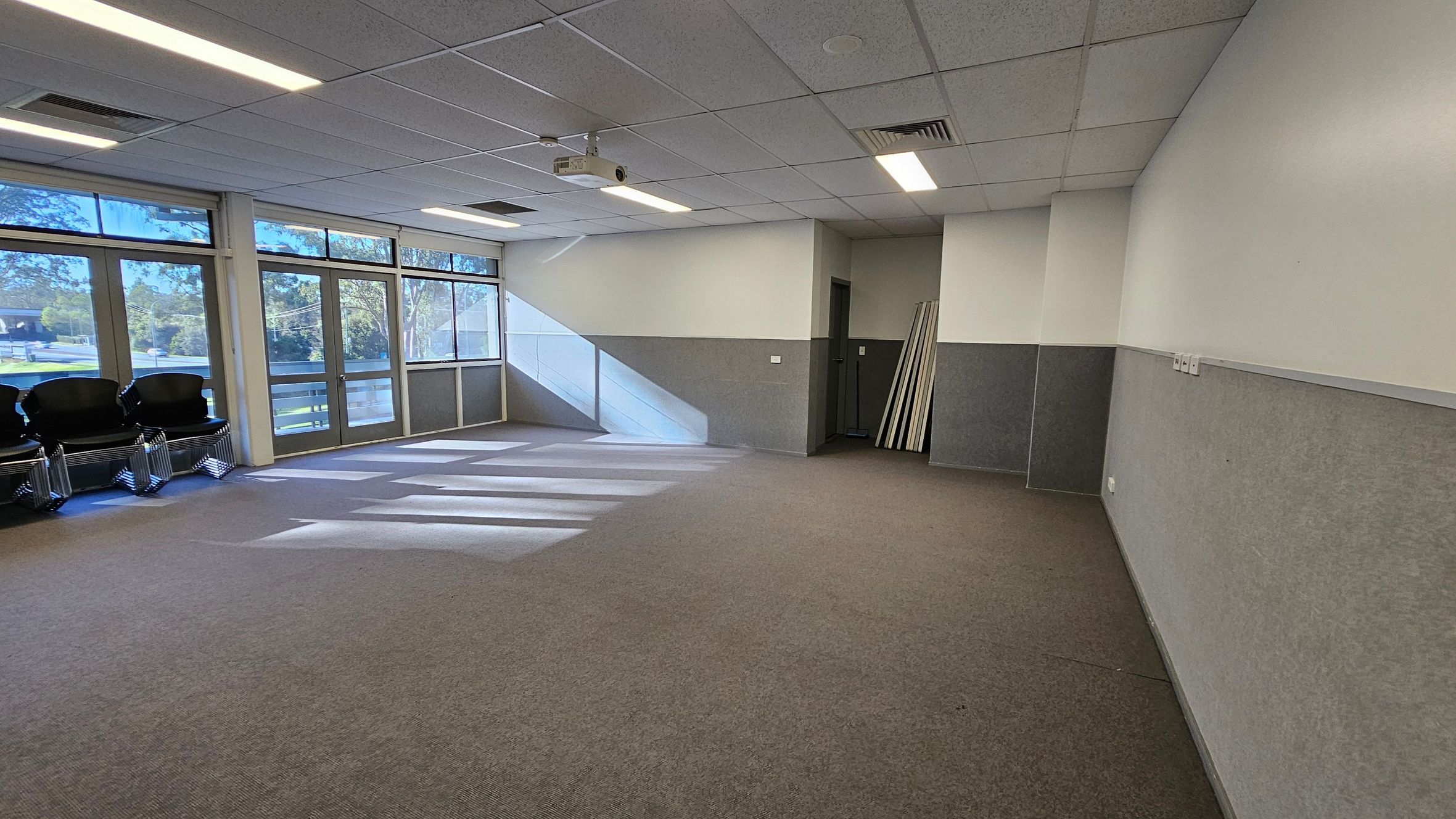A room with a grey and brown carpeted floor and a white panelled ceiling with fluorescent lights. On the left, there are large windows and glass doors allowing natural light to stream in, casting shadows on the floor. Several black chairs are stacked in the corner. On the right, the walls are painted in two tones, with a darker lower half. In the background, there is a small alcove with several long boards leaning against the wall. A ceiling-mounted projector is visible. A room with a grey and brown carpeted floor and a white panelled ceiling with fluorescent lights. On the left, there are large windows and glass doors allowing natural light to stream in, casting shadows on the floor. Several black chairs are stacked in the corner. On the right, the walls are painted in two tones, with a darker lower half. In the background, there is a small alcove with several long boards leaning against the wall. A ceiling-mounted projector is visible.