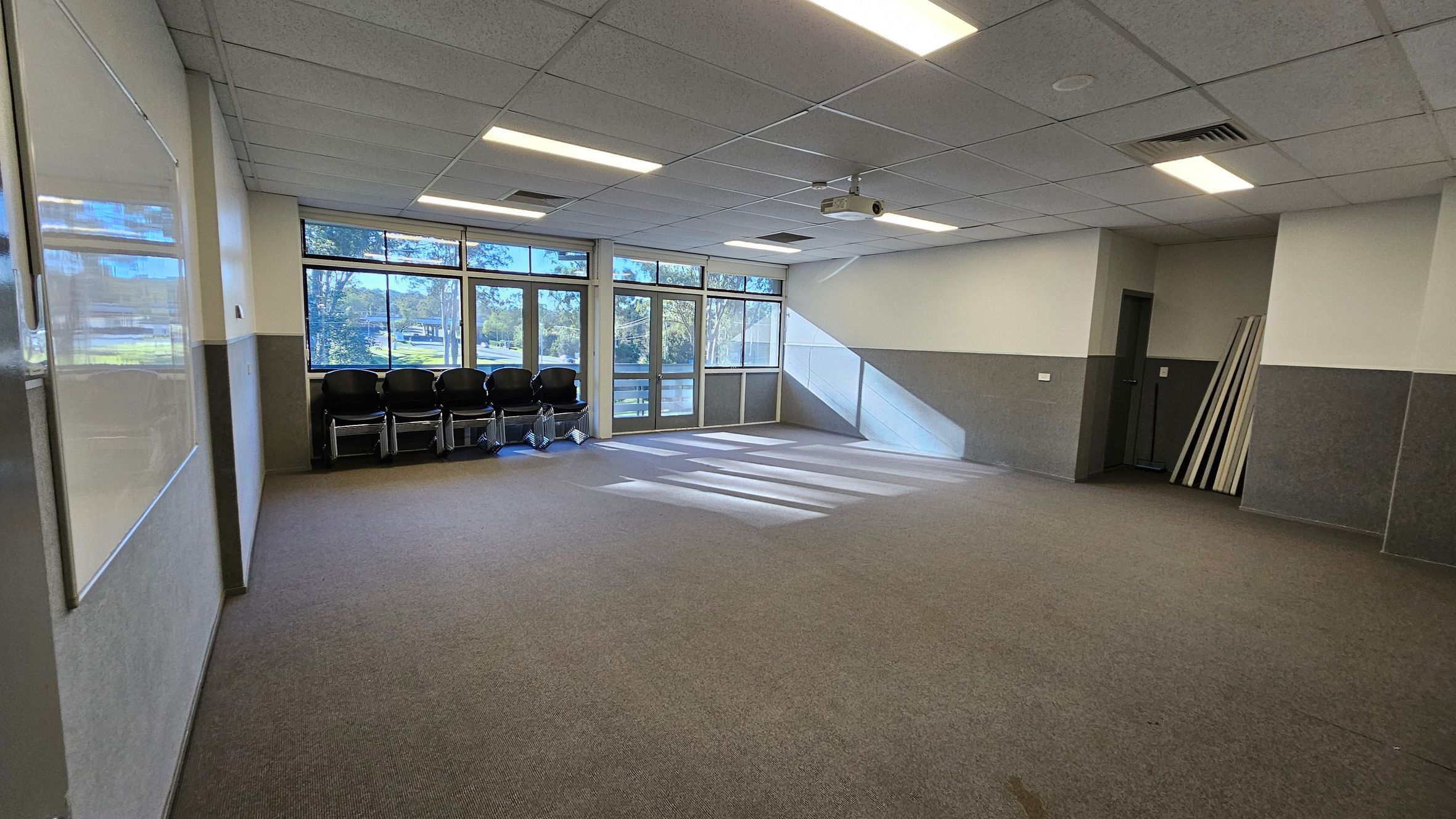 A spacious room with a grey and brown carpeted floor. On the left, there's a whiteboard on the wall. The back wall features large windows and glass doors, allowing natural light to stream in, casting shadows on the floor. Several stacked chairs are positioned against the window. On the right, a small alcove contains a door and some stacked panels. The ceiling is fitted with fluorescent lights and a projector. A spacious room with a grey and brown carpeted floor. On the left, there's a whiteboard on the wall. The back wall features large windows and glass doors, allowing natural light to stream in, casting shadows on the floor. Several stacked chairs are positioned against the window. On the right, a small alcove contains a door and some stacked panels. The ceiling is fitted with fluorescent lights and a projector.