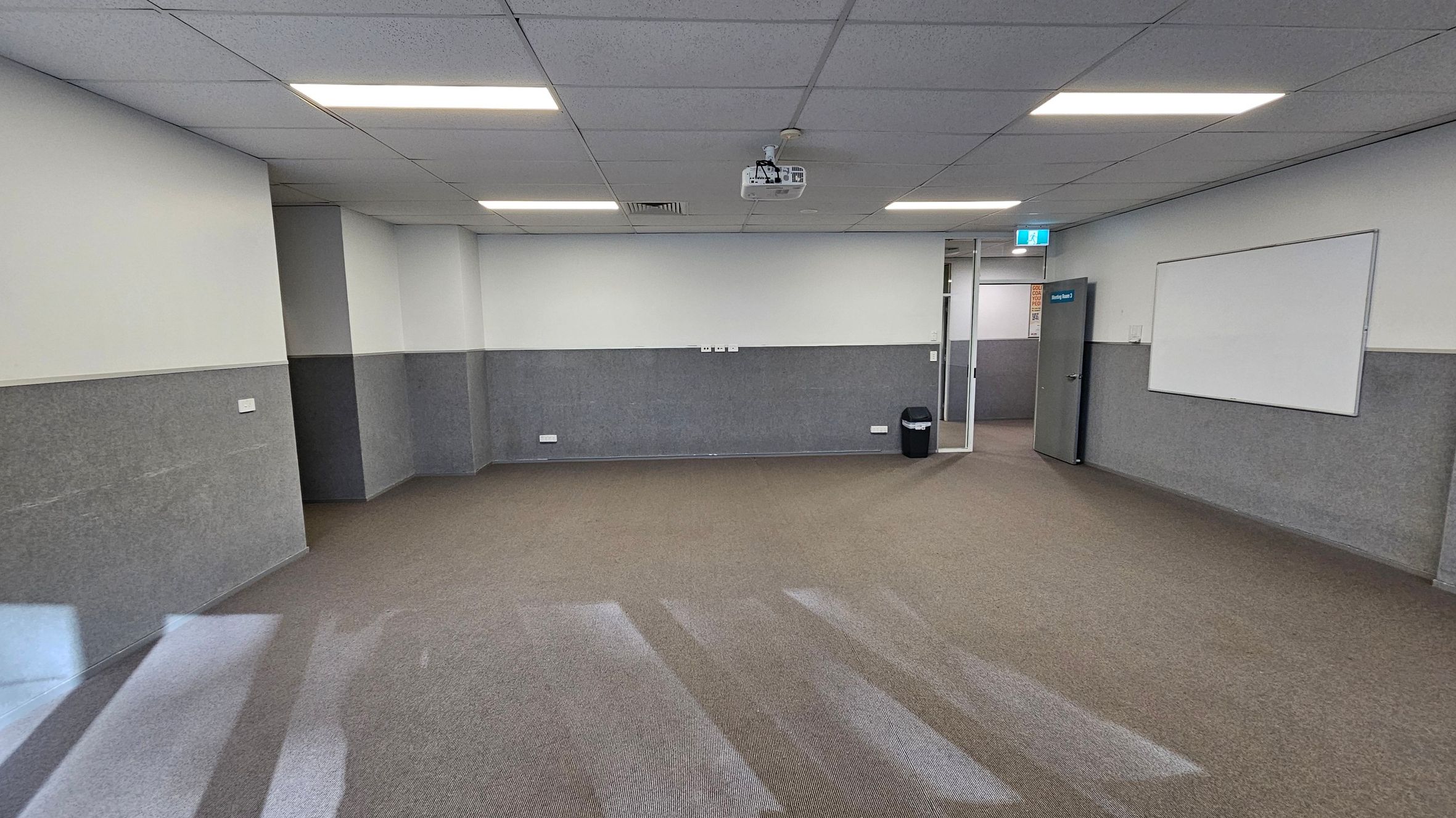 A room with grey and white walls, featuring a whiteboard on the right wall. The floor is covered with brown and grey carpet. A ceiling-mounted projector is in the centre, with fluorescent lights overhead. A door with a blue exit sign is on the right, next to a black bin. The ceiling is composed of grey tiles. A room with grey and white walls, featuring a whiteboard on the right wall. The floor is covered with brown and grey carpet. A ceiling-mounted projector is in the centre, with fluorescent lights overhead. A door with a blue exit sign is on the right, next to a black bin. The ceiling is composed of grey tiles.