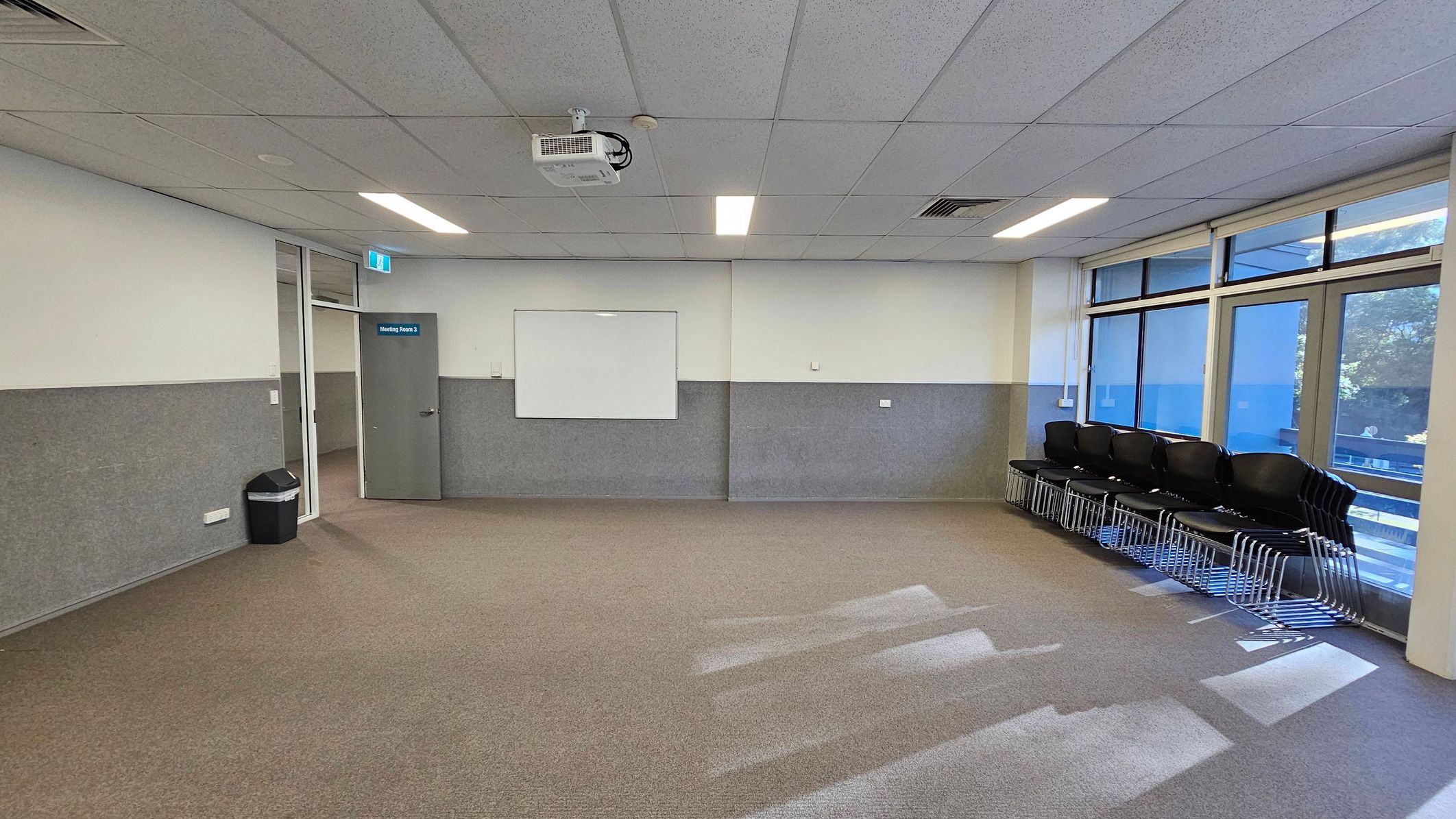 A meeting room with a brown carpeted floor and a white ceiling with fluorescent lights. On the left, there's a grey door labelled "Meeting Room 3" and a black bin. In the centre, a whiteboard is mounted on the wall. On the right, a stack of black chairs is placed against the wall, near large windows that let in natural light. A ceiling-mounted projector is visible above. A meeting room with a brown carpeted floor and a white ceiling with fluorescent lights. On the left, there's a grey door labelled "Meeting Room 3" and a black bin. In the centre, a whiteboard is mounted on the wall. On the right, a stack of black chairs is placed against the wall, near large windows that let in natural light. A ceiling-mounted projector is visible above.