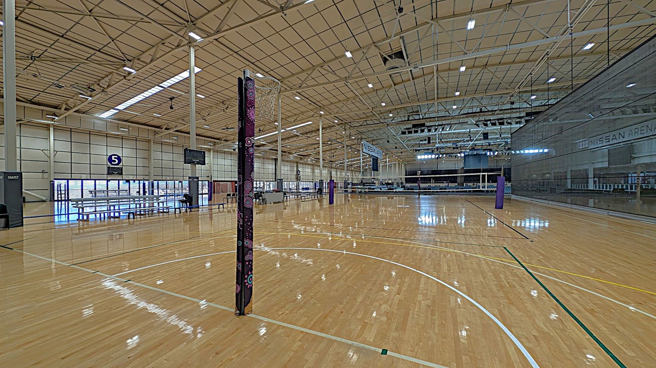 A spacious indoor sports arena with a polished wooden floor marked for netball. In the centre, a netball post with a protective cover stands prominently. On the left, there are rows of benches and a scoreboard. The ceiling is high with exposed beams and lighting fixtures. On the right, a large glass wall with "Nissan Arena" written on it. The background features more seating and additional netball posts.