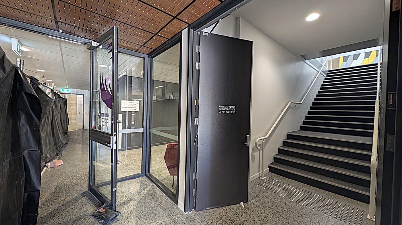 A hallway with a polished, speckled floor and a ceiling with recessed lighting. On the left, a long corridor with closed doors and signs on the wall. In the middle, a seating area with a green sofa and two red chairs against a wall with green foliage. On the right, a glass door leads to another area with covered items and an exit with glass doors showing an outdoor view.