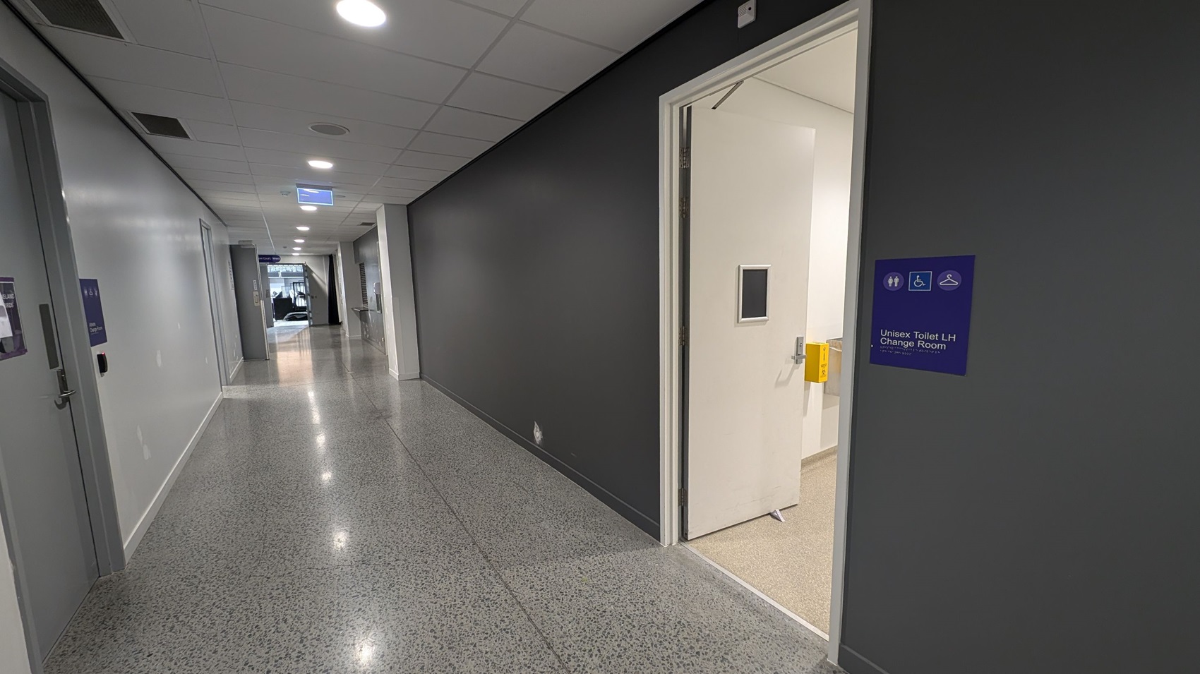 A corridor with a polished, speckled floor and a white ceiling with recessed lights. On the right, a door is open, leading to a unisex toilet and change room, indicated by a blue sign. The walls are painted grey. On the left, there are closed doors and a sign hanging from the ceiling. At the end of the corridor, there is a glass entrance or exit with natural light coming through.