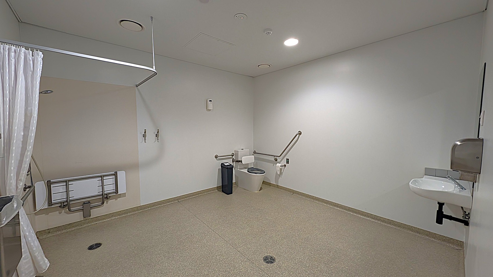 A spacious, accessible bathroom with a green speckled floor and white walls. On the right, there's a wall-mounted sink with a soap dispenser above it. In the centre, a toilet with support rails on both sides and a bin nearby. On the left, a folded wall-mounted changing table and a shower curtain rail. The ceiling is white with recessed lighting and ventilation.