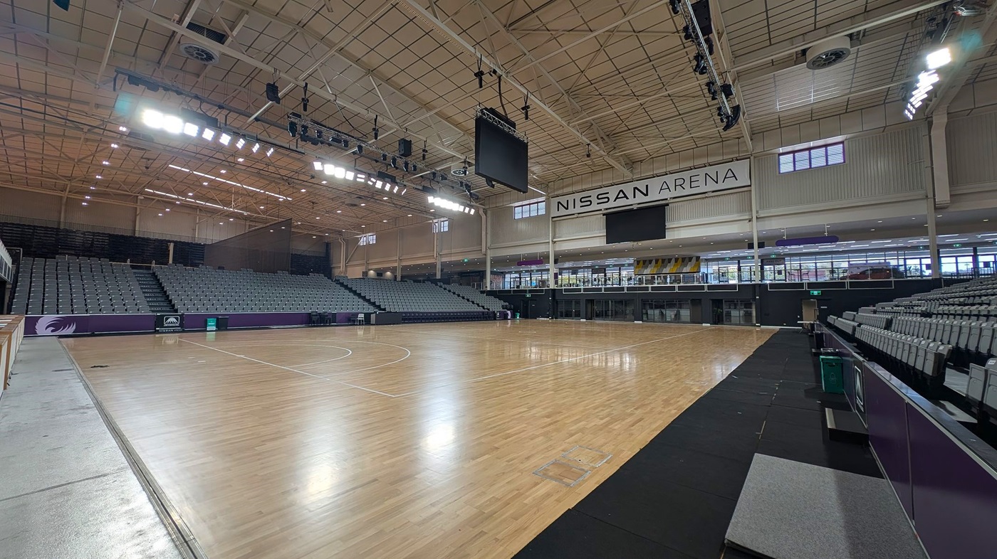 A large indoor sports arena with a polished wooden floor. On the left, there are tiered seating areas with grey seats. The right side also features similar seating. In the centre, a large scoreboard hangs from the ceiling. The ceiling is high with exposed beams and lighting fixtures. The back wall displays the name "Nissan Arena" above a row of windows and doors. The arena is well-lit, and the space is open and spacious.
