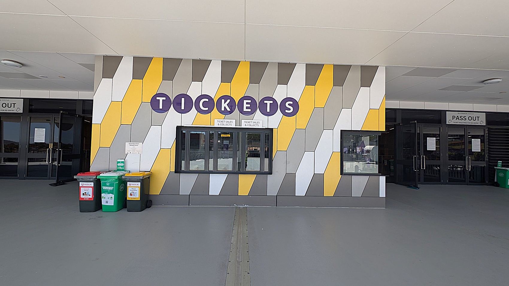 A ticket booth with a colourful geometric wall pattern featuring yellow, grey, and white shapes. The word "TICKETS" is displayed in large blue circles above the window. To the left, there are three bins with red, green, and yellow lids. On either side of the booth, there are glass doors with signs above them, one reading "PASS OUT". The floor is grey, and the ceiling is light-coloured with circular fixtures.