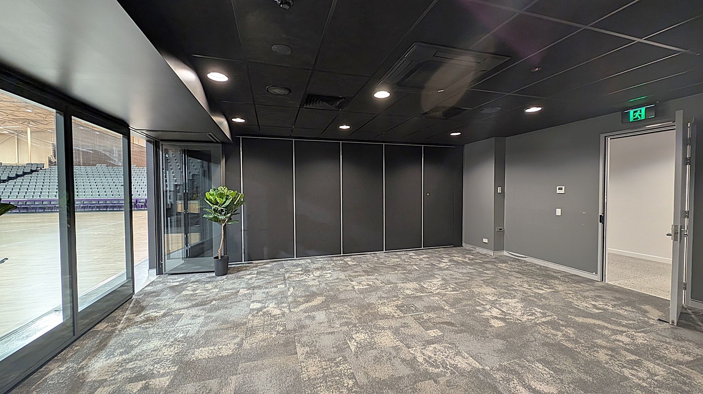 A room with a patterned carpet in shades of grey and green. On the left, large glass windows reveal a view of an adjacent area with rows of blue seats. A potted plant is near the windows. The back wall is dark with panelled sections. On the right, a door is open, leading to another room. The ceiling is dark with recessed lighting, and an illuminated exit sign is above the door.