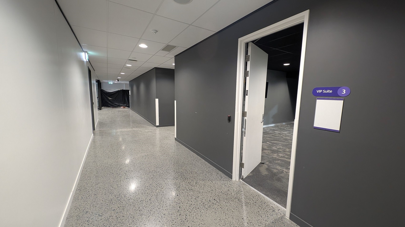 A corridor with polished speckled flooring and a white ceiling with recessed lights. On the right, a door is open, leading to a room labelled "VIP Suite 3" with a sign on the wall. The walls are painted grey. In the background, the corridor extends with a black curtain partially visible at the end.