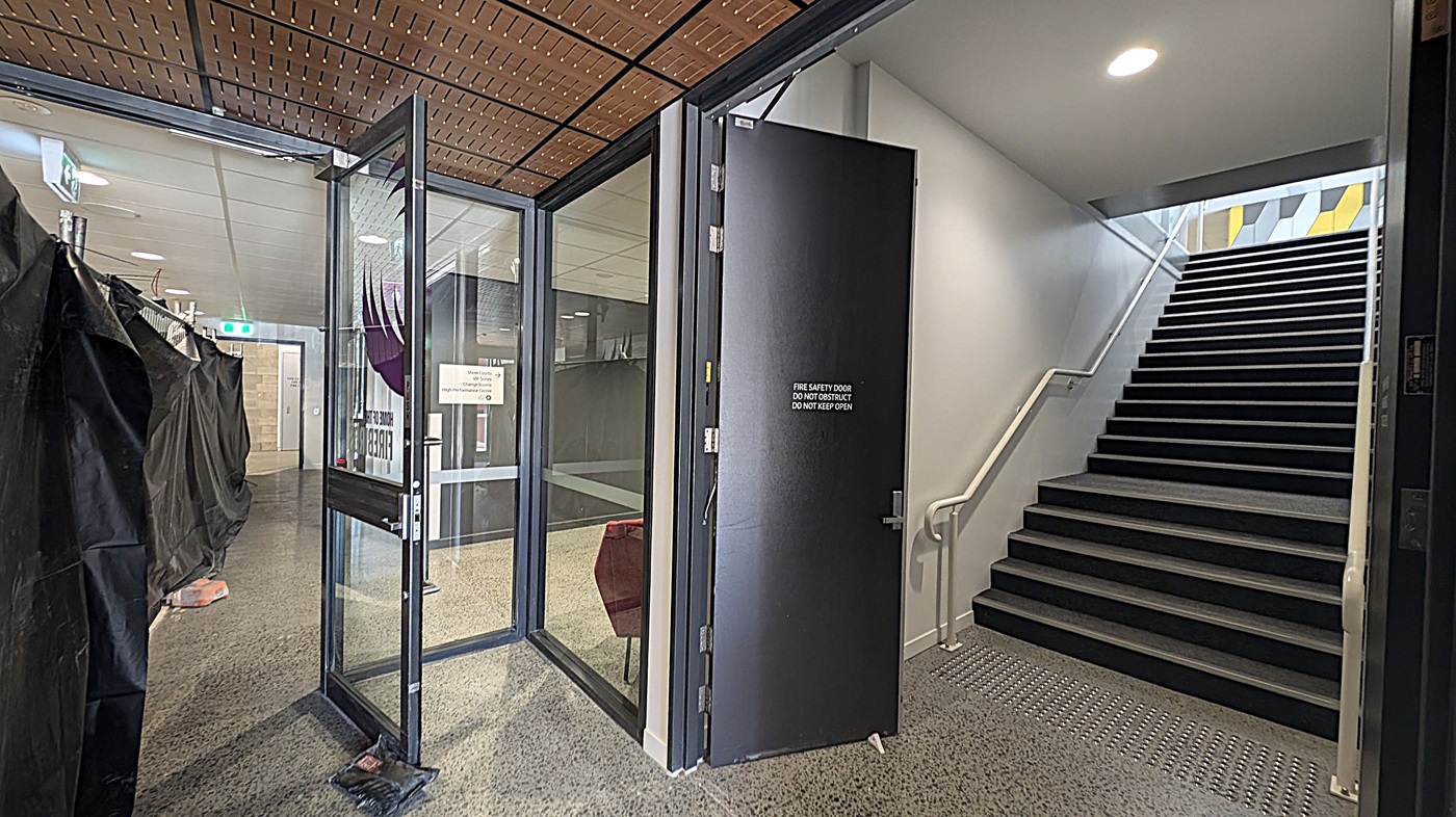 A hallway with a glass door on the left, partially open, leading to a corridor with black draped coverings. A sign on the door reads "FIRE SAFETY DOOR DO NOT OBSTRUCT DO NOT KEEP OPEN." On the right, a staircase with a white handrail ascends, with a patterned wall visible at the top. The ceiling is wooden with a grid pattern, and the floor is speckled. A round ceiling light illuminates the stairway area.