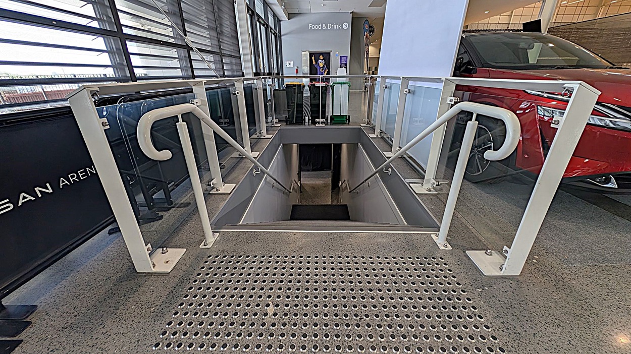 A staircase descends into a lower level, flanked by white railings and glass panels. The floor is speckled with a textured metal surface at the top of the stairs. To the right, a red car is partially visible. In the background, a sign reads "Food & Drink" with bins and a poster nearby. Large windows with horizontal blinds are on the left, allowing natural light into the space.
