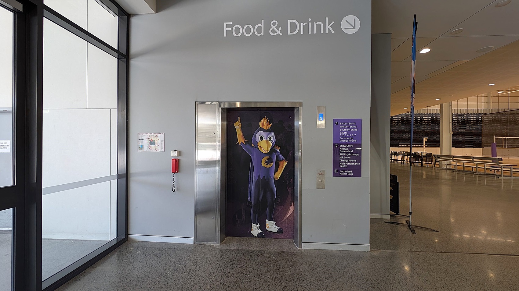 A hallway with a lift in the centre featuring a cartoon bird character on the door. Above, the words "Food & Drink" are displayed. To the left, large glass windows and a wall-mounted fire extinguisher. On the right, a sign with directions and a glimpse into a seating area with benches and tables. The floor is polished concrete, and the ceiling has recessed lighting.