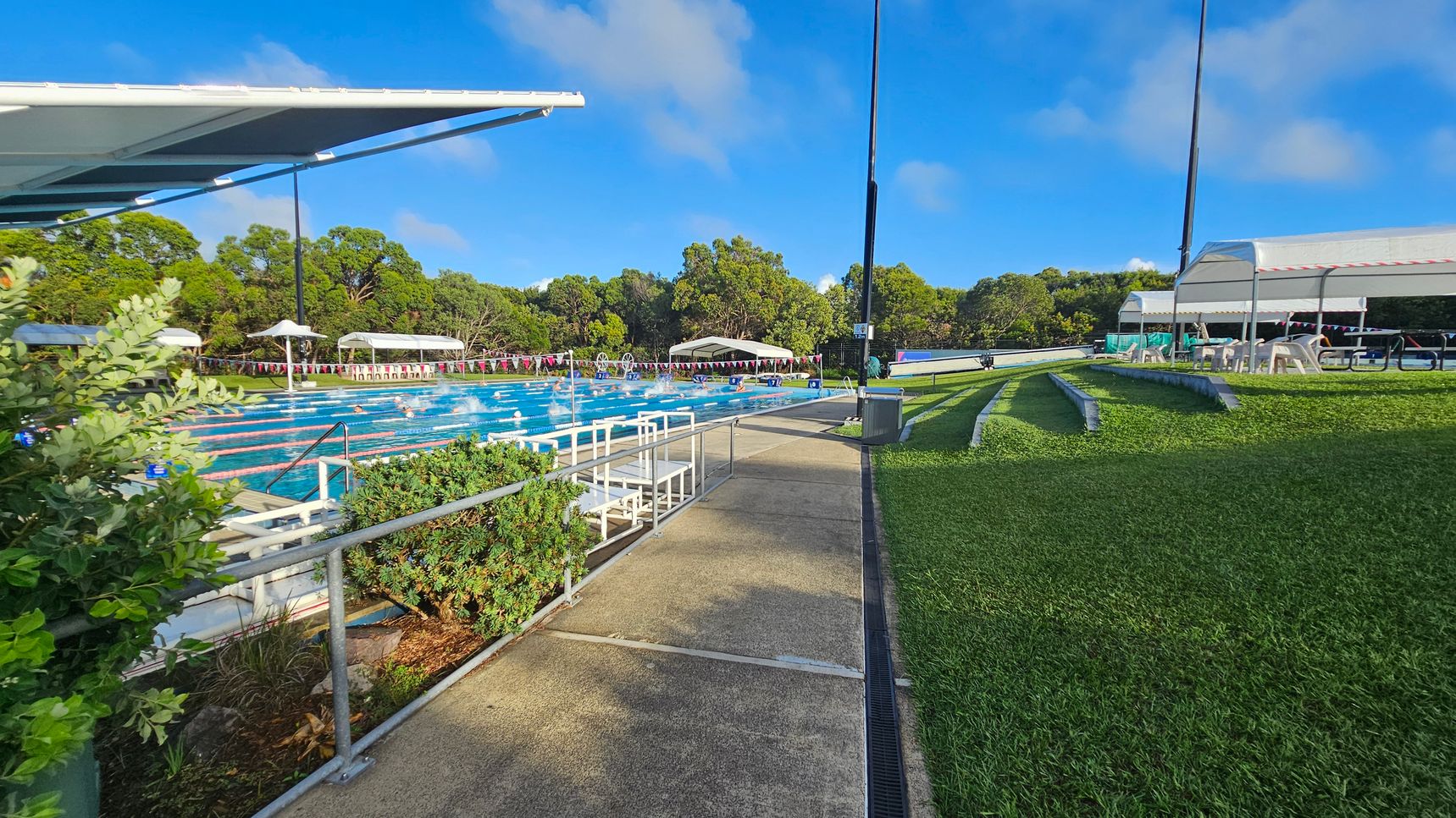 A swimming pool with multiple lanes is on the left, surrounded by greenery and shaded by a large canopy. Swimmers are visible in the water. On the right, there is a grassy area with tiered seating and a covered area with tables and chairs. The background features trees and a clear blue sky. A concrete path runs between the pool and the grassy area. A swimming pool with multiple lanes is on the left, surrounded by greenery and shaded by a large canopy. Swimmers are visible in the water. On the right, there is a grassy area with tiered seating and a covered area with tables and chairs. The background features trees and a clear blue sky. A concrete path runs between the pool and the grassy area.