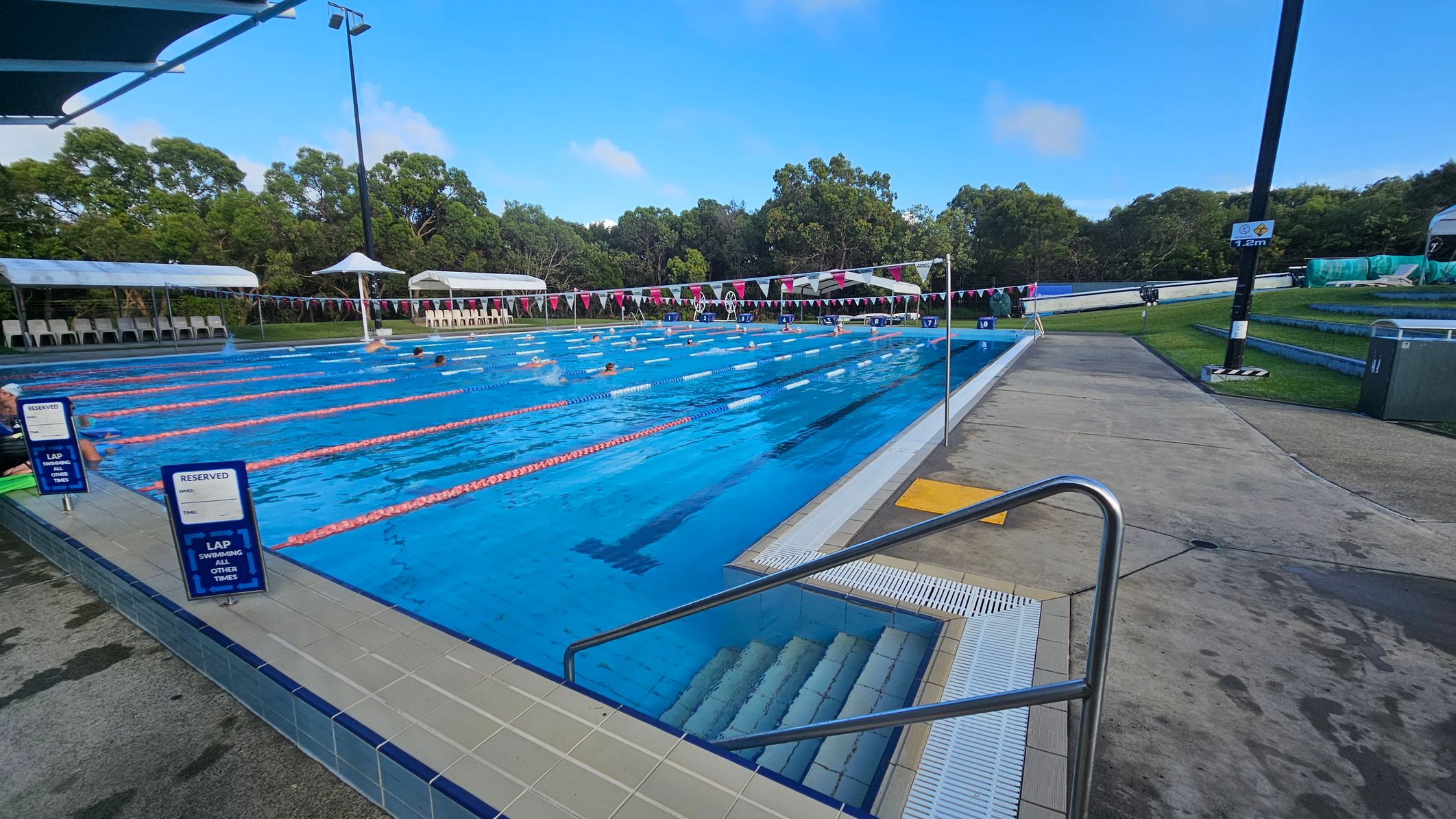 Outdoor swimming pool with multiple lanes, separated by orange lane dividers. Swimmers are in the water. On the left, there are signs indicating reserved lanes. The poolside is tiled, with steps leading into the water. In the background, there are white canopies and chairs, with trees beyond. On the right, a concrete path runs alongside the pool, with a grassy area and a tall light pole. Outdoor swimming pool with multiple lanes, separated by orange lane dividers. Swimmers are in the water. On the left, there are signs indicating reserved lanes. The poolside is tiled, with steps leading into the water. In the background, there are white canopies and chairs, with trees beyond. On the right, a concrete path runs alongside the pool, with a grassy area and a tall light pole.