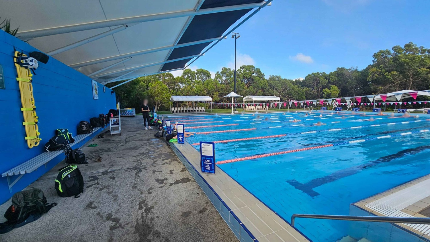 On the left, a blue wall with a yellow rescue board and several backpacks on a bench. The ground is wet. A white canopy covers the area. In the middle, a person stands near the edge of a swimming pool. On the right, a large outdoor pool with swimmers in designated lanes, marked by blue and red lane dividers. White signs indicate reserved lanes. In the background, trees and white seating areas with umbrellas are visible. On the left, a blue wall with a yellow rescue board and several backpacks on a bench. The ground is wet. A white canopy covers the area. In the middle, a person stands near the edge of a swimming pool. On the right, a large outdoor pool with swimmers in designated lanes, marked by blue and red lane dividers. White signs indicate reserved lanes. In the background, trees and white seating areas with umbrellas are visible.