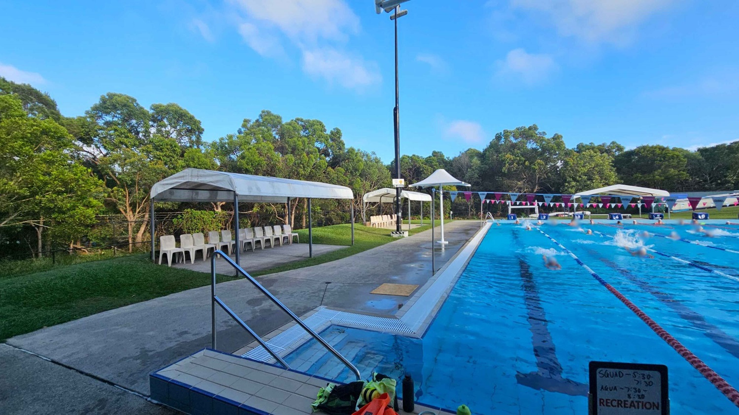 On the left, there are white plastic chairs under a canopy, with green trees in the background. In the middle, a concrete path runs alongside a swimming pool. The pool is on the right, with swimmers in lanes and colourful flags strung above. A sign with a schedule is visible in the foreground. The poolside area is wet, and there are some items like towels and bottles near the pool's edge. On the left, there are white plastic chairs under a canopy, with green trees in the background. In the middle, a concrete path runs alongside a swimming pool. The pool is on the right, with swimmers in lanes and colourful flags strung above. A sign with a schedule is visible in the foreground. The poolside area is wet, and there are some items like towels and bottles near the pool's edge.