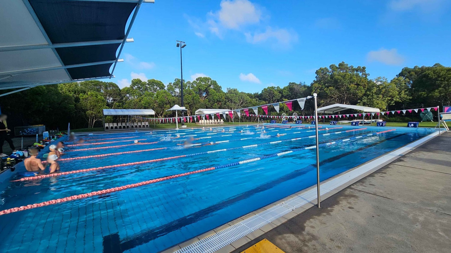 An outdoor swimming pool with multiple lanes, separated by pink lane dividers. Swimmers are in the water, with some resting at the edge on the left. A large canopy provides shade on the left side. In the background, there are white tents and chairs along the poolside, with trees and greenery beyond. The pool deck is concrete with a wet surface. Triangular flags are strung across the pool. An outdoor swimming pool with multiple lanes, separated by pink lane dividers. Swimmers are in the water, with some resting at the edge on the left. A large canopy provides shade on the left side. In the background, there are white tents and chairs along the poolside, with trees and greenery beyond. The pool deck is concrete with a wet surface. Triangular flags are strung across the pool.