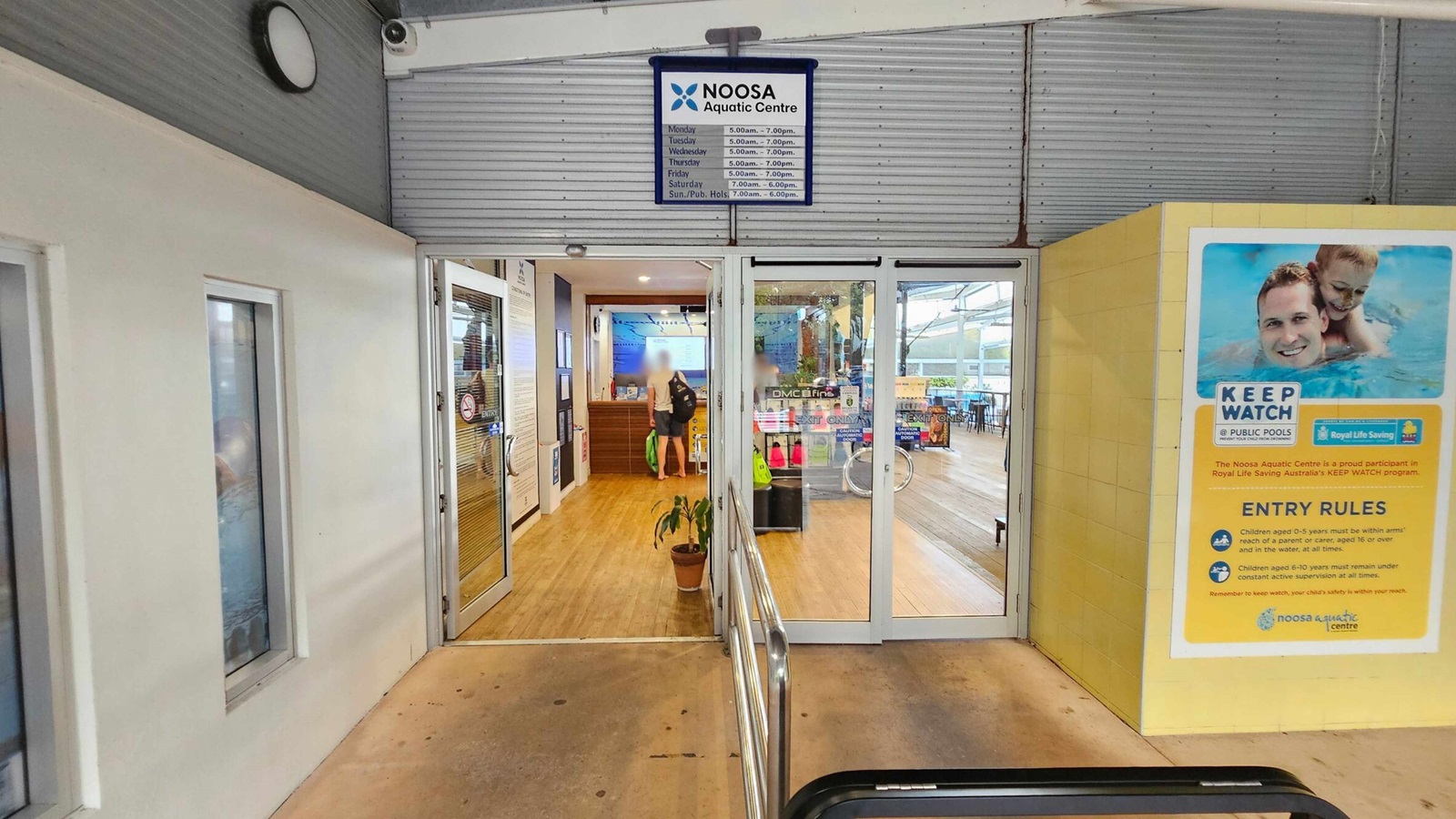 Entrance to the Noosa Aquatic Centre with glass doors in the centre. Above the doors, a sign displays the centre's name and opening hours. To the right, a large poster outlines pool safety rules with an image of a man and child swimming. Inside, a person stands near a wooden reception desk, and various swimming gear is visible. The floor is wooden, and a potted plant is placed near the entrance. The ceiling is light-coloured with a clock and security camera. Entrance to the Noosa Aquatic Centre with glass doors in the centre. Above the doors, a sign displays the centre's name and opening hours. To the right, a large poster outlines pool safety rules with an image of a man and child swimming. Inside, a person stands near a wooden reception desk, and various swimming gear is visible. The floor is wooden, and a potted plant is placed near the entrance. The ceiling is light-coloured with a clock and security camera.