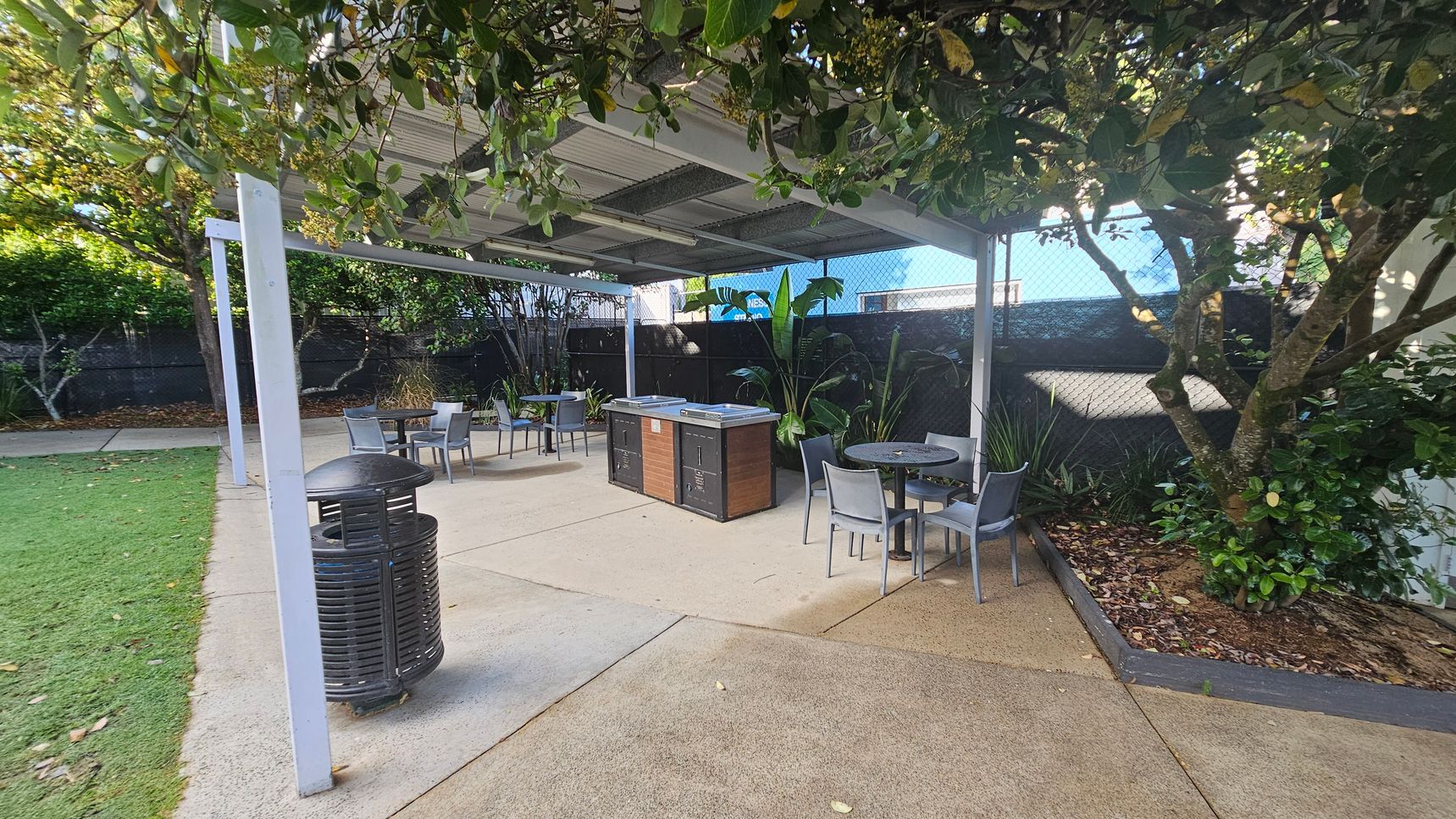 A covered outdoor area with a metal roof and white support beams. In the centre, there are two barbecue units and several round tables with grey chairs. The ground is a mix of concrete and grass. On the right, a tree with dense foliage provides shade, and there are various plants along the fence in the background. A black rubbish bin is on the left near the grass. A covered outdoor area with a metal roof and white support beams. In the centre, there are two barbecue units and several round tables with grey chairs. The ground is a mix of concrete and grass. On the right, a tree with dense foliage provides shade, and there are various plants along the fence in the background. A black rubbish bin is on the left near the grass.