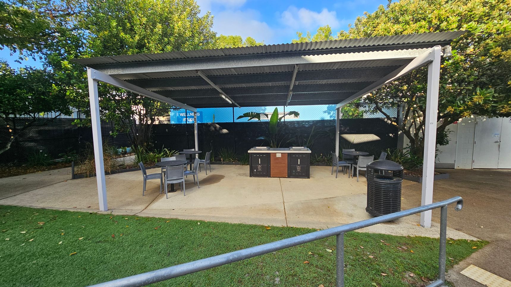 A covered outdoor area with a corrugated metal roof supported by white metal posts. In the centre, there are two barbecue grills with a wooden panel in between. Surrounding the grills are several grey tables and chairs. A black rubbish bin is on the right. The ground is a mix of concrete and artificial grass. In the background, there are trees and a blue sign partially visible, reading "WELLNESS STUDIO". A covered outdoor area with a corrugated metal roof supported by white metal posts. In the centre, there are two barbecue grills with a wooden panel in between. Surrounding the grills are several grey tables and chairs. A black rubbish bin is on the right. The ground is a mix of concrete and artificial grass. In the background, there are trees and a blue sign partially visible, reading "WELLNESS STUDIO".