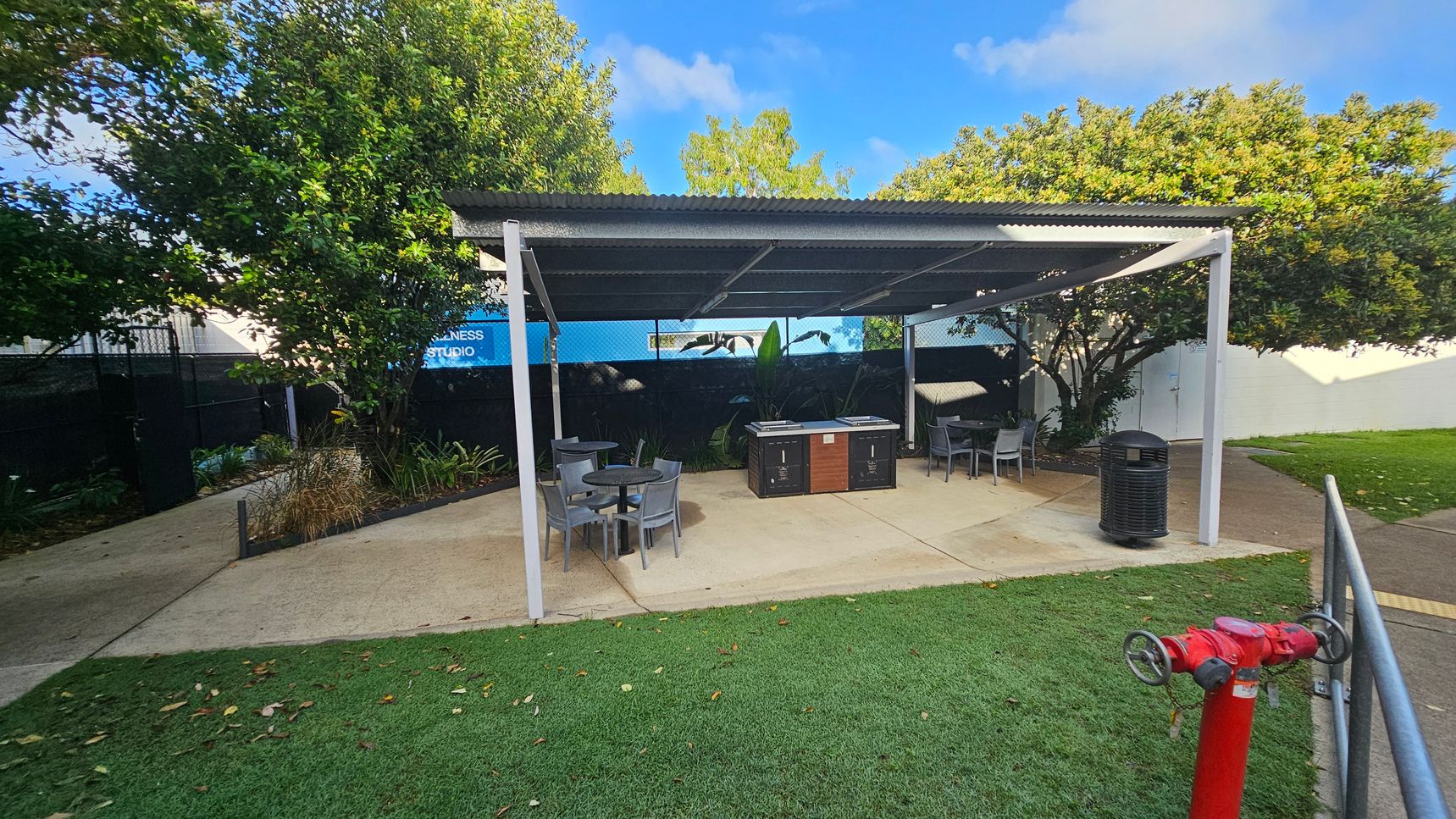 A covered outdoor area with a corrugated metal roof. Underneath, there are several tables and chairs on a concrete floor. To the right, a bin is visible. In the foreground, a red fire hydrant is on a grassy area. The background features lush green trees and a sign partially visible with the words "WELLNESS STUDIO." A pathway runs alongside the grass, leading towards the trees. A covered outdoor area with a corrugated metal roof. Underneath, there are several tables and chairs on a concrete floor. To the right, a bin is visible. In the foreground, a red fire hydrant is on a grassy area. The background features lush green trees and a sign partially visible with the words "WELLNESS STUDIO." A pathway runs alongside the grass, leading towards the trees.