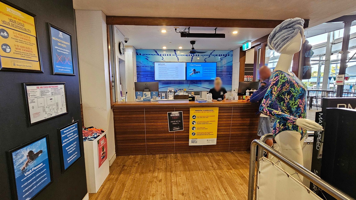 A café with a wooden interior. On the right, a barista is preparing drinks behind a counter with a display of pastries and a coffee machine. Shelves hold coffee supplies and a sign for coconut water. In the centre, a TV screen is mounted above the counter. On the left, an ice cream freezer with "Peters" branding is next to a display of clothing and accessories. The floor is wooden, and the ceiling is white with recessed lighting. A café with a wooden interior. On the right, a barista is preparing drinks behind a counter with a display of pastries and a coffee machine. Shelves hold coffee supplies and a sign for coconut water. In the centre, a TV screen is mounted above the counter. On the left, an ice cream freezer with "Peters" branding is next to a display of clothing and accessories. The floor is wooden, and the ceiling is white with recessed lighting.
