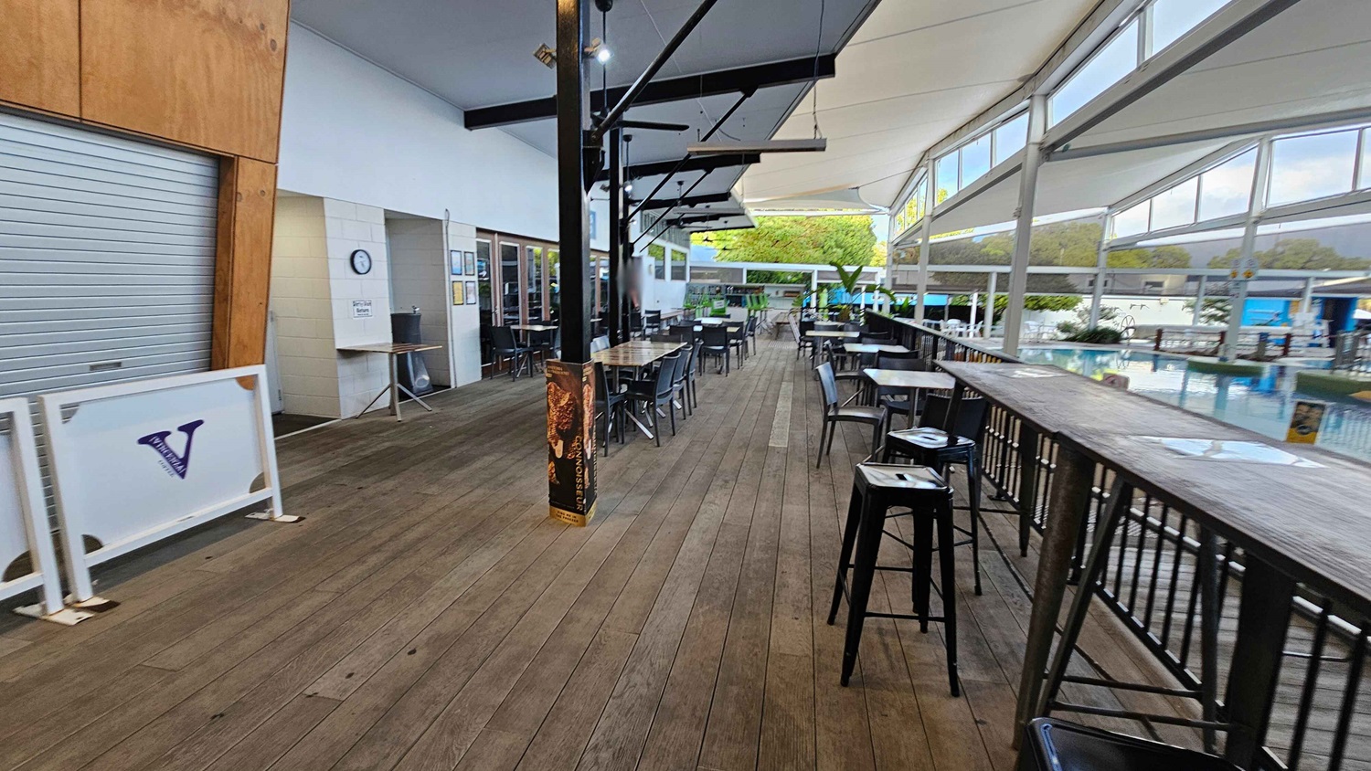 A spacious outdoor seating area with wooden flooring. On the left, there's a closed metal shutter and a sign with a "V" logo. In the middle, several tables and chairs are arranged under a canopy with black support beams. On the right, a long wooden bar with high stools overlooks a pool area. Large windows and a white ceiling allow natural light to fill the space. Greenery is visible in the background near the pool. A spacious outdoor seating area with wooden flooring. On the left, there's a closed metal shutter and a sign with a "V" logo. In the middle, several tables and chairs are arranged under a canopy with black support beams. On the right, a long wooden bar with high stools overlooks a pool area. Large windows and a white ceiling allow natural light to fill the space. Greenery is visible in the background near the pool.