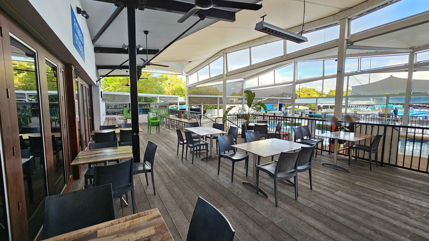 Outdoor seating area with wooden tables and black chairs on a wooden deck. On the left, glass doors reflect the surroundings. In the middle, tables are arranged in rows, with some stacked green chairs in the background. The right side features a railing overlooking a pool area with people visible in the distance. The ceiling is covered with a white canopy, and large windows allow natural light to fill the space. Outdoor seating area with wooden tables and black chairs on a wooden deck. On the left, glass doors reflect the surroundings. In the middle, tables are arranged in rows, with some stacked green chairs in the background. The right side features a railing overlooking a pool area with people visible in the distance. The ceiling is covered with a white canopy, and large windows allow natural light to fill the space.