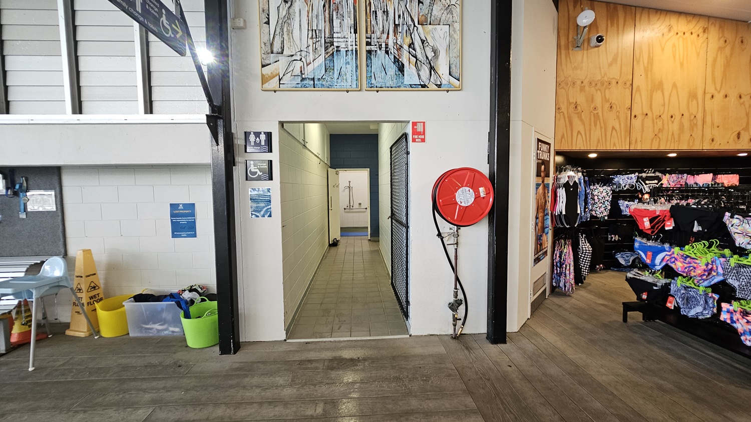A corridor with tiled flooring leads to a changing area. On the left, there are chairs, wet floor signs, and storage bins. On the right, a red fire hose reel is mounted on the wall, and a shop area displays swimwear on racks. The walls are white with signage for accessible toilets. Above the corridor entrance, there is a colourful artwork. The ceiling is wooden on the right side. A corridor with tiled flooring leads to a changing area. On the left, there are chairs, wet floor signs, and storage bins. On the right, a red fire hose reel is mounted on the wall, and a shop area displays swimwear on racks. The walls are white with signage for accessible toilets. Above the corridor entrance, there is a colourful artwork. The ceiling is wooden on the right side.