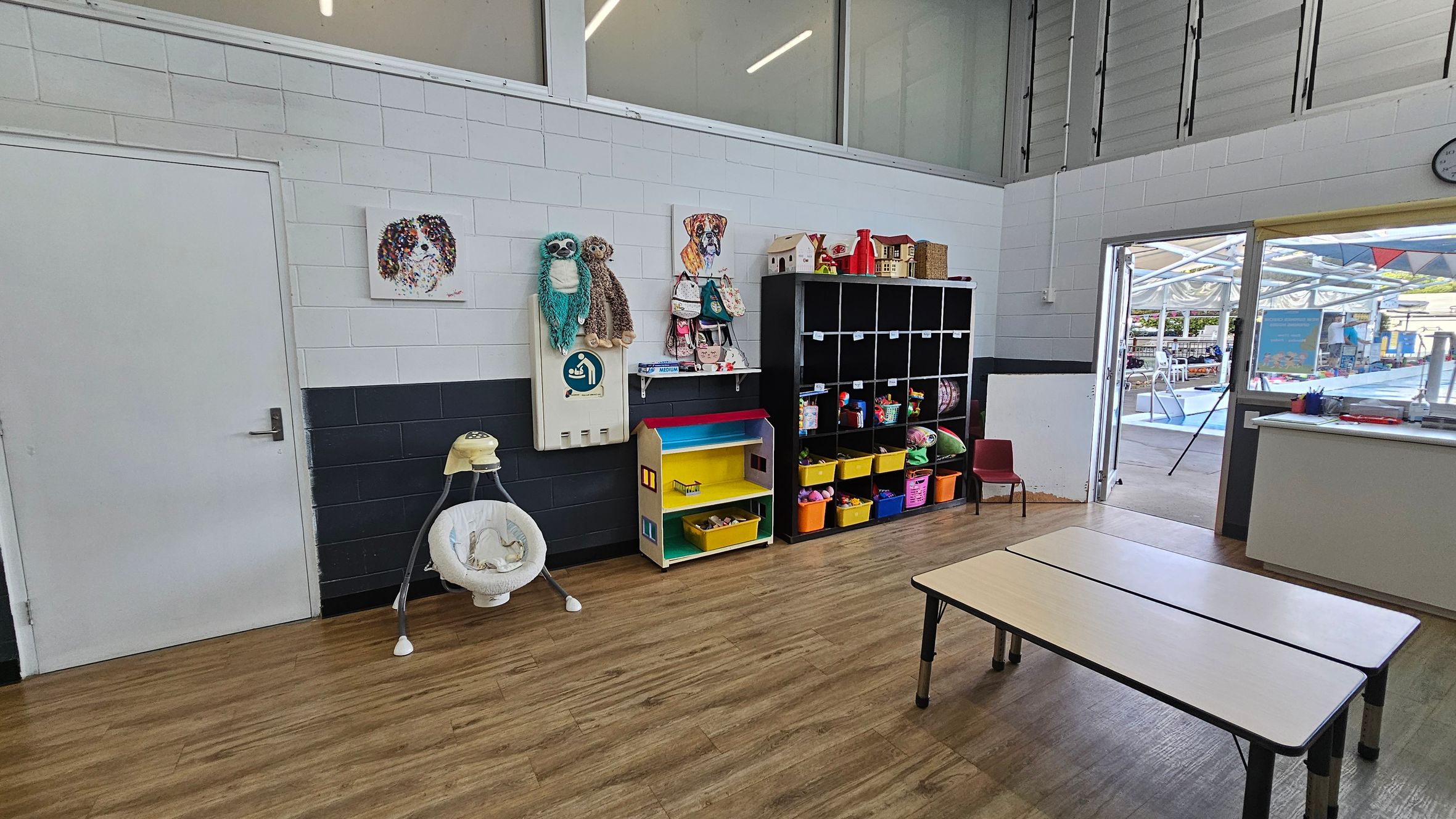 A classroom with a wooden floor and white ceiling. On the left, there's a white door and a baby swing. The wall has a painting of a dog and two plush toys hanging. In the middle, a colourful shelf with yellow bins and a black cubby unit filled with toys. On the right, two white tables and a small red chair. A door leads outside to a covered area with a pool. A clock is visible on the wall near the door. A classroom with a wooden floor and white ceiling. On the left, there's a white door and a baby swing. The wall has a painting of a dog and two plush toys hanging. In the middle, a colourful shelf with yellow bins and a black cubby unit filled with toys. On the right, two white tables and a small red chair. A door leads outside to a covered area with a pool. A clock is visible on the wall near the door.