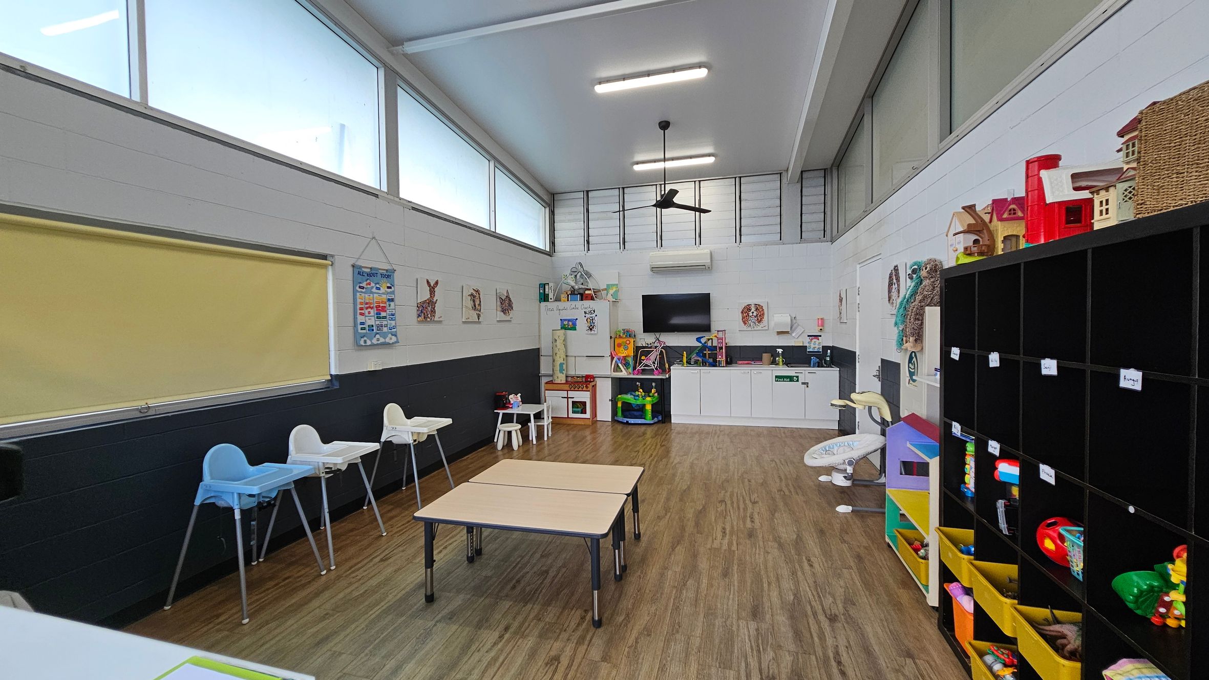 A brightly lit playroom with large windows near the ceiling. On the left, there are high chairs and a yellow blind. In the centre, two small tables are placed on a wooden floor. The back wall features a kitchenette with a TV above it, and various toys and educational materials. On the right, a black cubby shelf holds toys and baskets. The ceiling has fluorescent lights and a ceiling fan. A brightly lit playroom with large windows near the ceiling. On the left, there are high chairs and a yellow blind. In the centre, two small tables are placed on a wooden floor. The back wall features a kitchenette with a TV above it, and various toys and educational materials. On the right, a black cubby shelf holds toys and baskets. The ceiling has fluorescent lights and a ceiling fan.