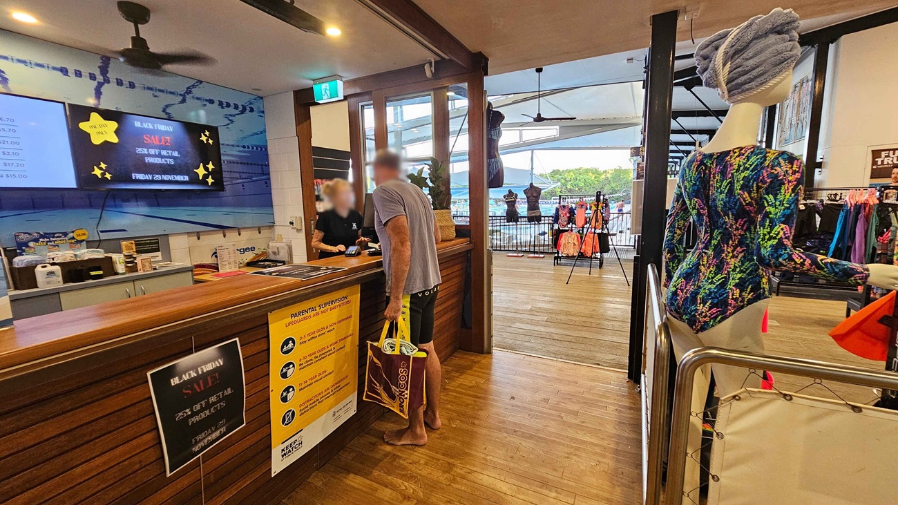 To the left, a wooden counter features a sales sign and various pricing boards above it, with a staff member assisting a customer. On the right, a mannequin dressed in a colourful swimsuit stands beside a display of swimwear and accessories. In the background, large windows allow light to filter through, revealing more swimwear on racks. The floor has a polished wooden surface, and the ceiling is plain with several light fixtures and ceiling fans visible To the left, a wooden counter features a sales sign and various pricing boards above it, with a staff member assisting a customer. On the right, a mannequin dressed in a colourful swimsuit stands beside a display of swimwear and accessories. In the background, large windows allow light to filter through, revealing more swimwear on racks. The floor has a polished wooden surface, and the ceiling is plain with several light fixtures and ceiling fans visible