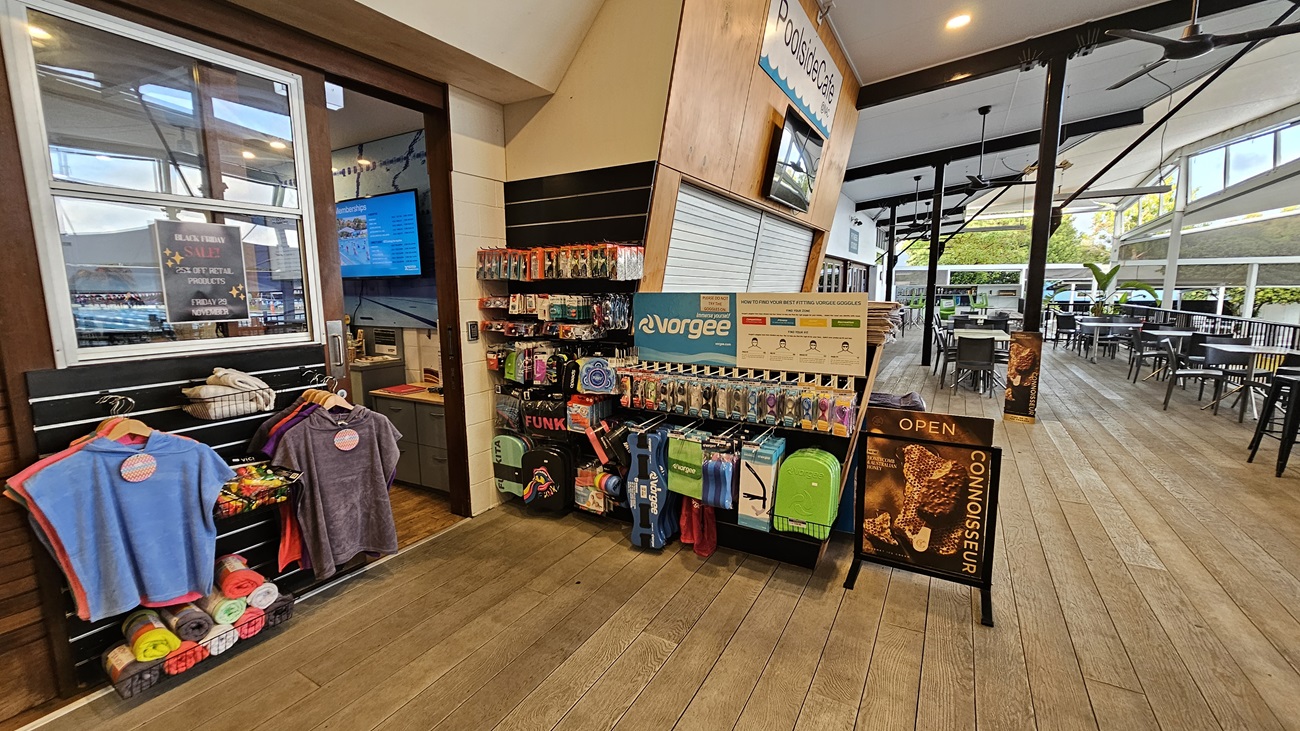 To the left, a display features a variety of colourful towels hung on racks, with a sign indicating the café nearby. Next to it, a wall-mounted screen shows information, and the counter displays various products for sale. On the right, a range of swimming accessories and bags is neatly organised on shelves, with an “Open” sign visible. The wooden floor runs throughout the area, while the ceiling has exposed beams and ceiling fans, contributing to an open and airy atmosphere. To the left, a display features a variety of colourful towels hung on racks, with a sign indicating the café nearby. Next to it, a wall-mounted screen shows information, and the counter displays various products for sale. On the right, a range of swimming accessories and bags is neatly organised on shelves, with an “Open” sign visible. The wooden floor runs throughout the area, while the ceiling has exposed beams and ceiling fans, contributing to an open and airy atmosphere.