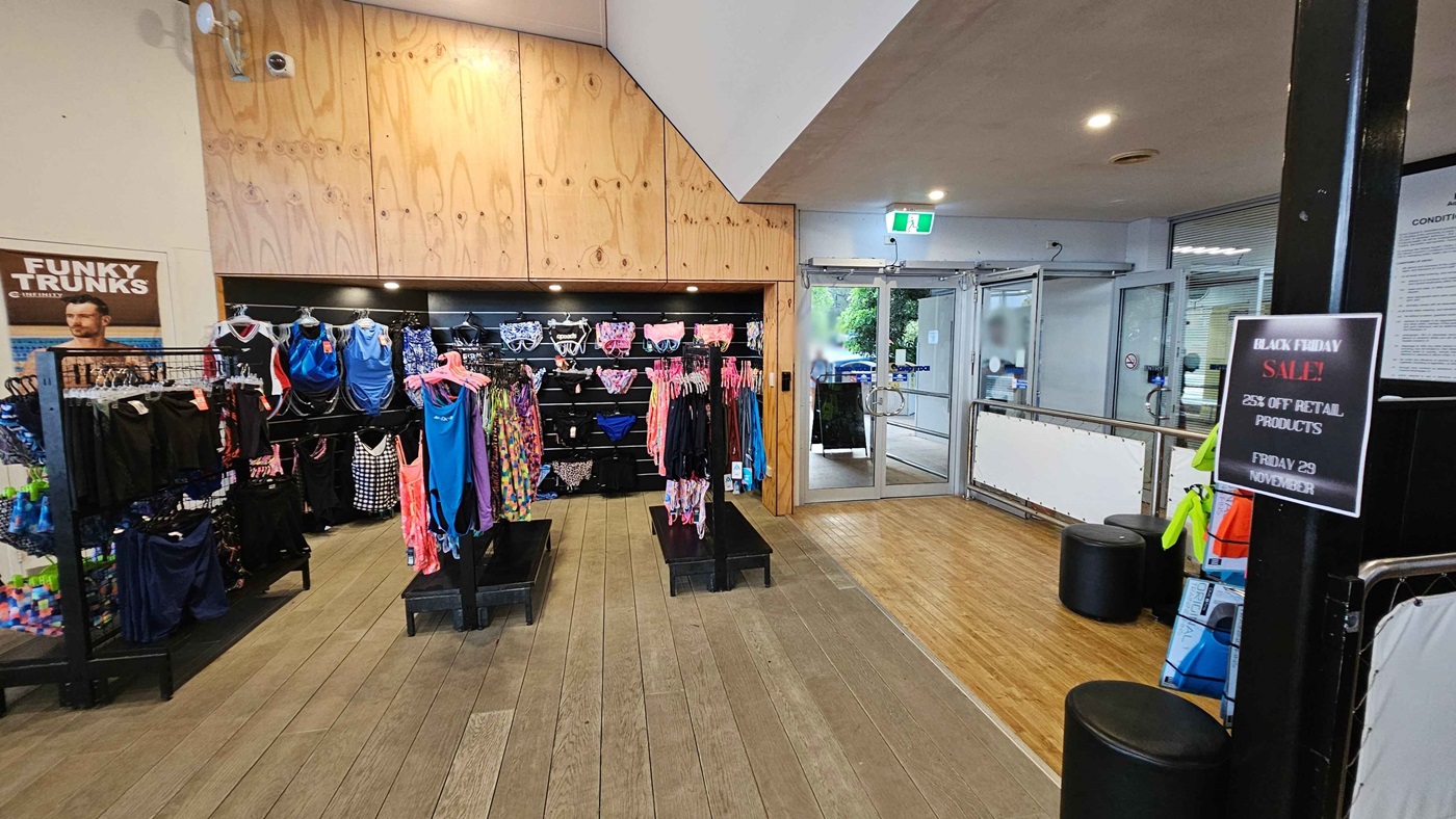On the left, there are racks displaying various swimwear items, including brightly coloured swimsuits and trunks, with a promotional poster for “Funky Trunks” nearby. To the right, a large glass door leads outside, and additional swim accessories are visible on a nearby display, alongside a "Black Friday Sale" sign advertising discounts. The floor features light wooden planks that contribute to a spacious feel, and the ceiling is neutral, with basic lighting fixtures overhead. On the left, there are racks displaying various swimwear items, including brightly coloured swimsuits and trunks, with a promotional poster for “Funky Trunks” nearby. To the right, a large glass door leads outside, and additional swim accessories are visible on a nearby display, alongside a "Black Friday Sale" sign advertising discounts. The floor features light wooden planks that contribute to a spacious feel, and the ceiling is neutral, with basic lighting fixtures overhead.