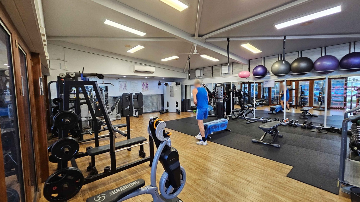 A gym with wooden flooring and a high ceiling with fluorescent lights. On the left, there are weight machines and a Kranking machine. In the centre, a person in blue athletic wear stands near a bench and exercise balls. On the right, a wall of mirrors reflects the gym equipment, including dumbbells and kettlebells. Large exercise balls are stored above the mirrors. The background features windows and more gym equipment. A gym with wooden flooring and a high ceiling with fluorescent lights. On the left, there are weight machines and a Kranking machine. In the centre, a person in blue athletic wear stands near a bench and exercise balls. On the right, a wall of mirrors reflects the gym equipment, including dumbbells and kettlebells. Large exercise balls are stored above the mirrors. The background features windows and more gym equipment.