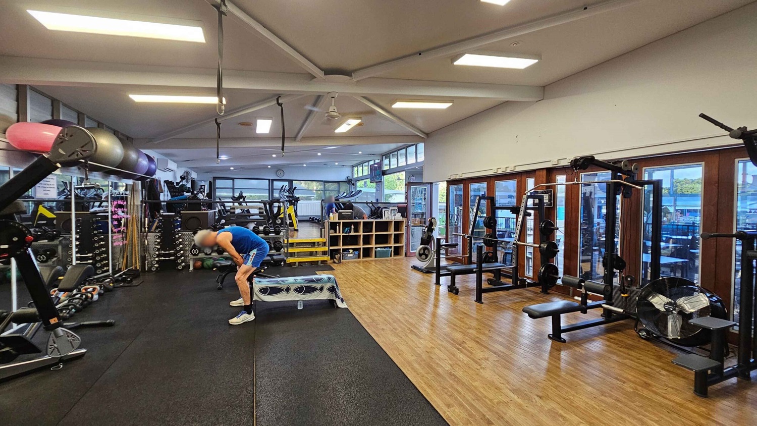 A gym interior with various exercise equipment. On the left, there are exercise balls and weights stacked on shelves. In the centre, a person in a blue tank top and shorts is bent over, resting on a bench. The floor is a mix of rubber and wood. On the right, there are weight machines and a large fan. The background features windows and mirrors, with more gym equipment visible, including treadmills and stationary bikes. The ceiling has exposed beams and fluorescent lights. A gym interior with various exercise equipment. On the left, there are exercise balls and weights stacked on shelves. In the centre, a person in a blue tank top and shorts is bent over, resting on a bench. The floor is a mix of rubber and wood. On the right, there are weight machines and a large fan. The background features windows and mirrors, with more gym equipment visible, including treadmills and stationary bikes. The ceiling has exposed beams and fluorescent lights.