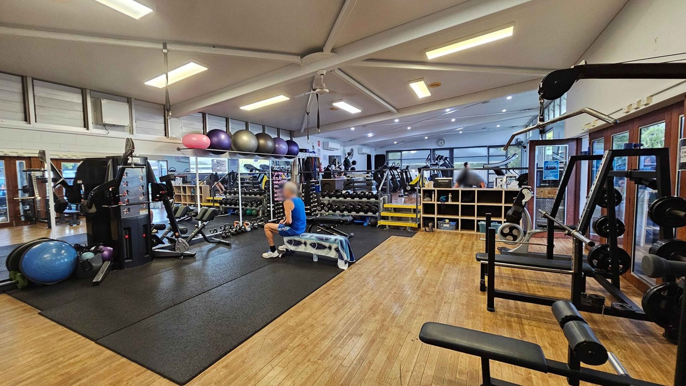 A gym interior with wooden flooring and a high, white ceiling with exposed beams. On the left, there are exercise balls and a multi-gym machine on a black mat. In the centre, a person in a blue shirt sits on a bench. Behind them, a wall of mirrors reflects rows of dumbbells and kettlebells. On the right, there is a weight rack and a cable machine. The background features treadmills and a clock on the wall. Large windows line the right side, allowing natural light in. A gym interior with wooden flooring and a high, white ceiling with exposed beams. On the left, there are exercise balls and a multi-gym machine on a black mat. In the centre, a person in a blue shirt sits on a bench. Behind them, a wall of mirrors reflects rows of dumbbells and kettlebells. On the right, there is a weight rack and a cable machine. The background features treadmills and a clock on the wall. Large windows line the right side, allowing natural light in.