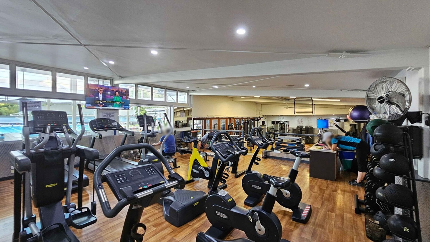 A gym with wooden flooring and a white ceiling. On the left, there are several elliptical machines facing a row of windows. A television is mounted above, displaying a news programme. In the middle, stationary bikes are lined up, with a few people exercising. On the right, a person leans on a desk near a rack of medicine balls. A large fan is mounted on the wall above. In the background, various weight machines and gym equipment are visible. A gym with wooden flooring and a white ceiling. On the left, there are several elliptical machines facing a row of windows. A television is mounted above, displaying a news programme. In the middle, stationary bikes are lined up, with a few people exercising. On the right, a person leans on a desk near a rack of medicine balls. A large fan is mounted on the wall above. In the background, various weight machines and gym equipment are visible.