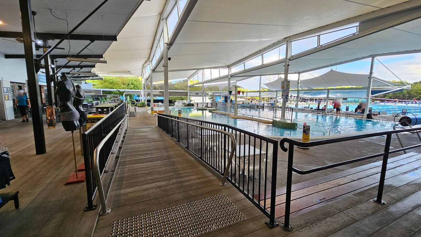 A spacious indoor swimming pool area with a wooden walkway on the left, featuring black mannequin torsos displaying swimwear. The walkway is bordered by a black metal railing. On the right, the pool is visible with people sitting and swimming. The ceiling is covered with large white fabric panels, and there are large windows allowing natural light. In the background, there are trees and more poolside seating. A spacious indoor swimming pool area with a wooden walkway on the left, featuring black mannequin torsos displaying swimwear. The walkway is bordered by a black metal railing. On the right, the pool is visible with people sitting and swimming. The ceiling is covered with large white fabric panels, and there are large windows allowing natural light. In the background, there are trees and more poolside seating.