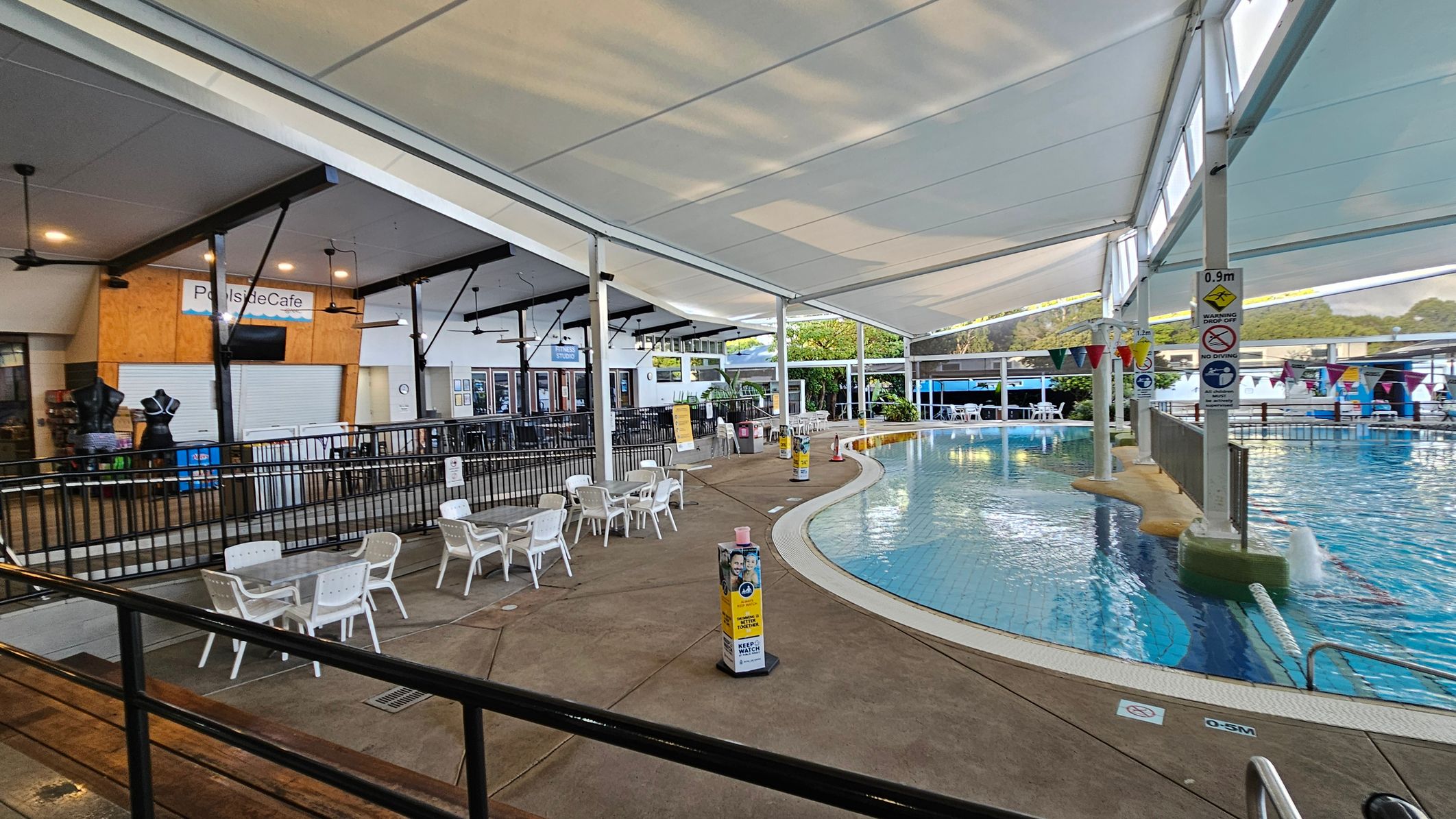 On the left, there is a closed café with a sign reading "Poolside Cafe" above it. Several ceiling fans are visible. In front of the café, there are white plastic tables and chairs on a concrete floor. On the right, a swimming pool with clear water is partially visible, surrounded by a fence. Colourful flags hang above the pool. The area is covered by a large white canopy, and there are safety signs near the pool. On the left, there is a closed café with a sign reading "Poolside Cafe" above it. Several ceiling fans are visible. In front of the café, there are white plastic tables and chairs on a concrete floor. On the right, a swimming pool with clear water is partially visible, surrounded by a fence. Colourful flags hang above the pool. The area is covered by a large white canopy, and there are safety signs near the pool.