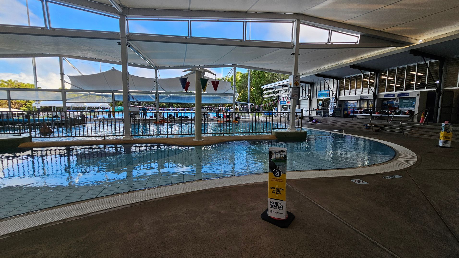 Indoor swimming pool area with a shallow pool in the foreground, surrounded by a tiled floor. A sign with safety instructions is visible near the pool's edge. In the background, there are larger pools with people swimming and lounging. The ceiling is covered with white fabric canopies, and large windows allow natural light to enter. On the right, there are steps leading to a seating area with informational boards and a few people sitting. Indoor swimming pool area with a shallow pool in the foreground, surrounded by a tiled floor. A sign with safety instructions is visible near the pool's edge. In the background, there are larger pools with people swimming and lounging. The ceiling is covered with white fabric canopies, and large windows allow natural light to enter. On the right, there are steps leading to a seating area with informational boards and a few people sitting.
