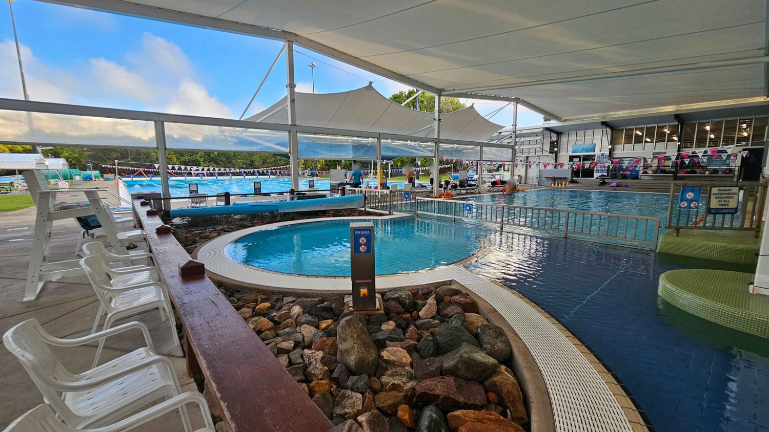 On the left, there are several white plastic chairs lined up along a wooden railing. In the middle, a circular spa pool is surrounded by a rock border and a sign with spa rules. To the right, a larger swimming pool is visible with people swimming and sitting on the edge. The area is covered by a large white canopy, and there are colourful flags strung above the pools. In the background, there is a grassy area and more pool lanes. On the left, there are several white plastic chairs lined up along a wooden railing. In the middle, a circular spa pool is surrounded by a rock border and a sign with spa rules. To the right, a larger swimming pool is visible with people swimming and sitting on the edge. The area is covered by a large white canopy, and there are colourful flags strung above the pools. In the background, there is a grassy area and more pool lanes.