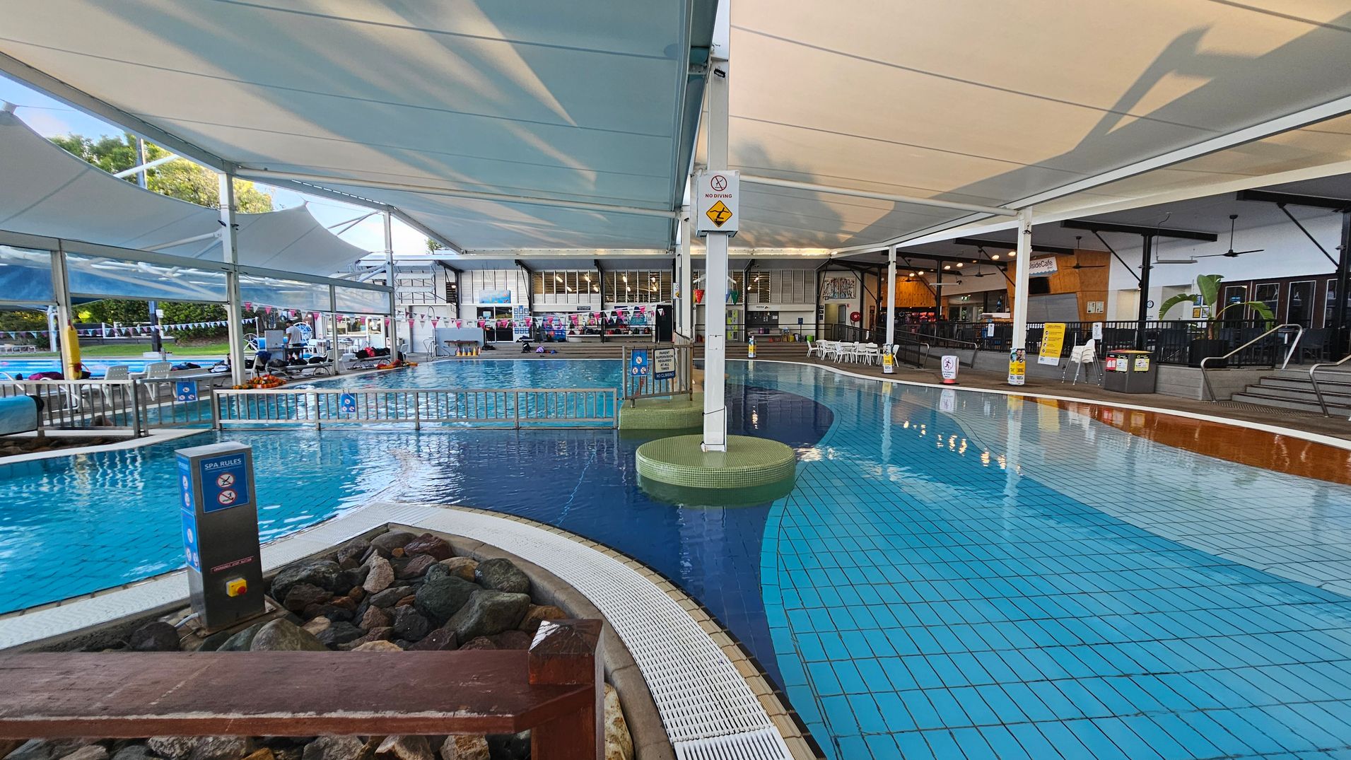 Indoor swimming pool area with a tiled floor and a curved pool. On the left, there are lounge chairs and a small fence separating the pool sections. A sign with spa rules is visible near a rocky area. The ceiling is covered with large white fabric canopies. On the right, there is a seating area with tables and chairs, and a café counter in the background. The area is well-lit with overhead lights, and there are signs and safety notices around the pool. Indoor swimming pool area with a tiled floor and a curved pool. On the left, there are lounge chairs and a small fence separating the pool sections. A sign with spa rules is visible near a rocky area. The ceiling is covered with large white fabric canopies. On the right, there is a seating area with tables and chairs, and a café counter in the background. The area is well-lit with overhead lights, and there are signs and safety notices around the pool.
