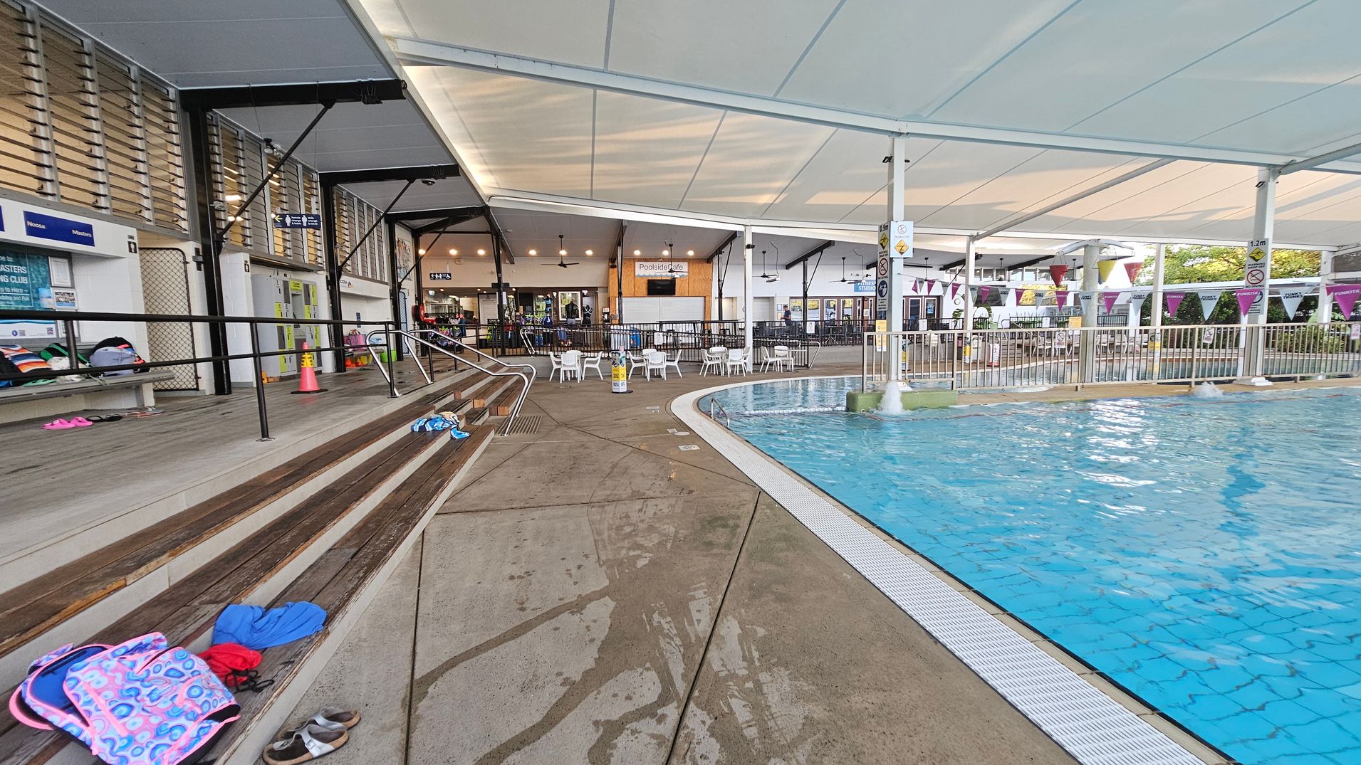 Indoor swimming pool area with a clear blue pool on the right. The pool is bordered by a white grate and surrounded by a wet concrete floor. On the left, there are wooden benches with various items like bags and shoes scattered on them. Above, a white ceiling with metal supports provides shelter. In the background, there are tables and chairs near a café area, with signs and a fence separating the seating from the pool. Indoor swimming pool area with a clear blue pool on the right. The pool is bordered by a white grate and surrounded by a wet concrete floor. On the left, there are wooden benches with various items like bags and shoes scattered on them. Above, a white ceiling with metal supports provides shelter. In the background, there are tables and chairs near a café area, with signs and a fence separating the seating from the pool.