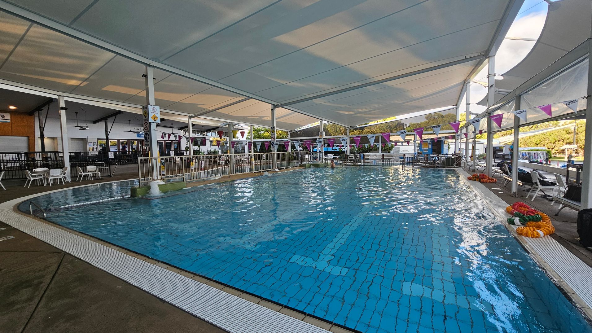 An indoor swimming pool with clear blue water, surrounded by a white tiled edge. On the left, there are white plastic chairs and tables near a black railing. The ceiling is covered with a large white canopy. On the right, colourful pool lane dividers are coiled on the ground, and more seating is visible. Bunting with purple and white flags hangs above the pool. In the background, there are more facilities and greenery outside. An indoor swimming pool with clear blue water, surrounded by a white tiled edge. On the left, there are white plastic chairs and tables near a black railing. The ceiling is covered with a large white canopy. On the right, colourful pool lane dividers are coiled on the ground, and more seating is visible. Bunting with purple and white flags hangs above the pool. In the background, there are more facilities and greenery outside.