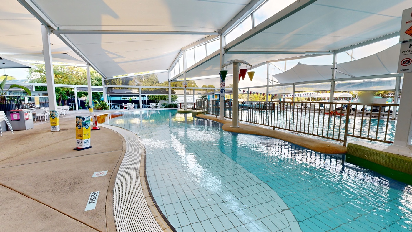 A tiled swimming pool curves through the centre, with light blue water reflecting overhead structures. On the left, there is a concrete path, chairs, tables, and a few bins. To the right, metal fencing runs along the pool edge. The foreground shows a shallow pool section, while the background includes greenery and shaded seating areas under white canopy roofs. The ceiling above is light and supported by metal beams. A tiled swimming pool curves through the centre, with light blue water reflecting overhead structures. On the left, there is a concrete path, chairs, tables, and a few bins. To the right, metal fencing runs along the pool edge. The foreground shows a shallow pool section, while the background includes greenery and shaded seating areas under white canopy roofs. The ceiling above is light and supported by metal beams.
