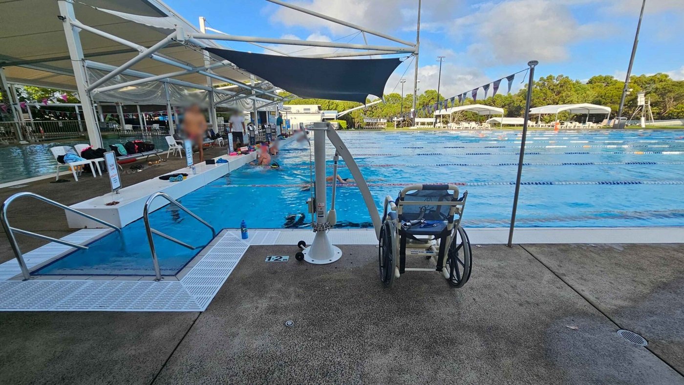 A swimming pool with lanes is in the centre, surrounded by a concrete deck. On the left, there are steps leading into the pool and a few people standing and sitting under a shaded area with chairs and bags. A wheelchair is positioned near a pool lift in the foreground. On the right, the pool extends with lane dividers, and in the background, there are trees and more shaded seating areas. The sky is partly cloudy. A swimming pool with lanes is in the centre, surrounded by a concrete deck. On the left, there are steps leading into the pool and a few people standing and sitting under a shaded area with chairs and bags. A wheelchair is positioned near a pool lift in the foreground. On the right, the pool extends with lane dividers, and in the background, there are trees and more shaded seating areas. The sky is partly cloudy.
