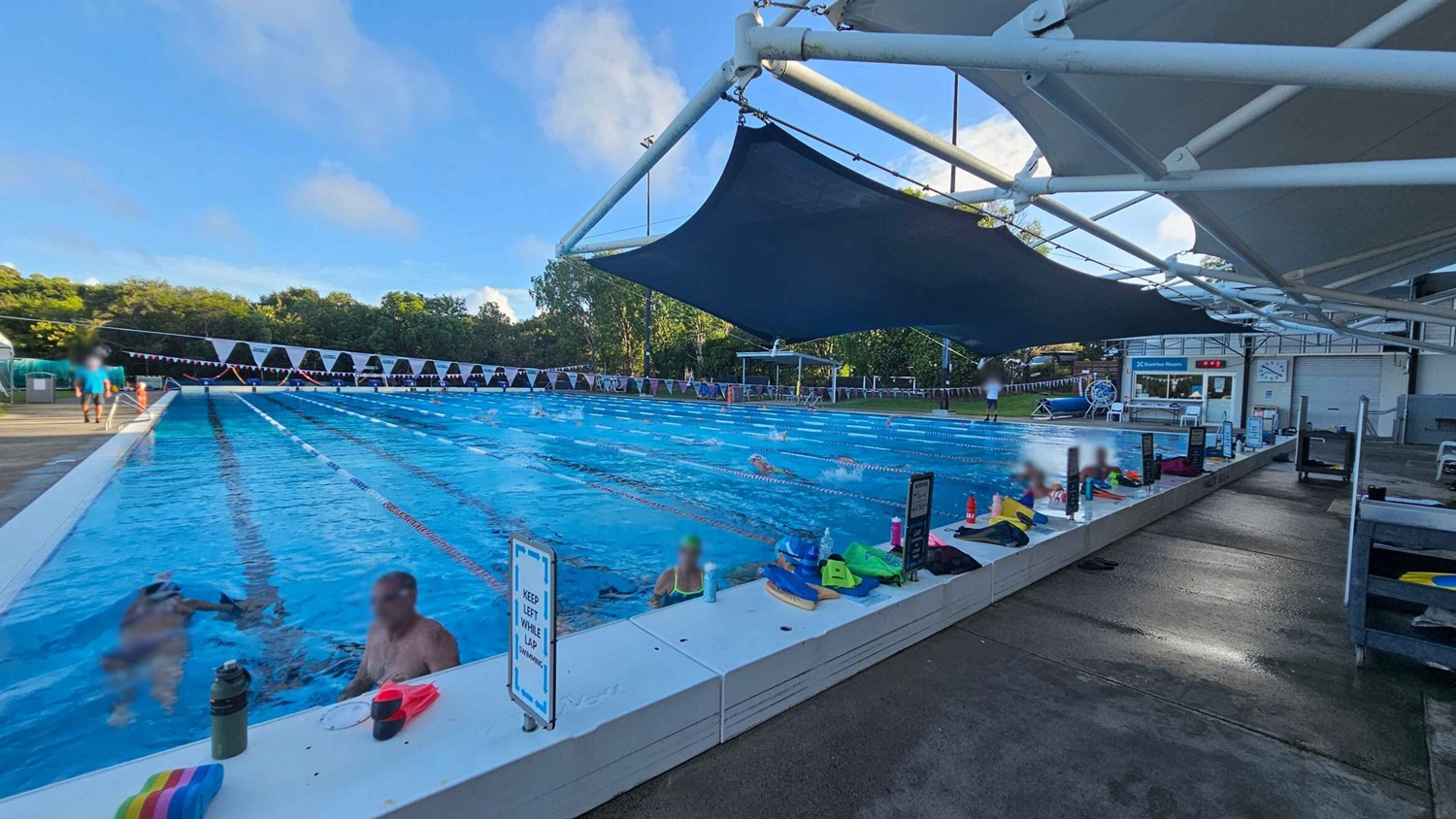 An outdoor swimming pool with multiple lanes, where several swimmers are practising. On the right, a shaded area with a white structure and dark blue fabric provides cover. The pool deck is lined with various swimming gear, water bottles, and signs. In the background, there are trees and a small building with a clock and a sign reading "Control Room." The pool is bordered by a white edge, and the deck is wet and brown. An outdoor swimming pool with multiple lanes, where several swimmers are practising. On the right, a shaded area with a white structure and dark blue fabric provides cover. The pool deck is lined with various swimming gear, water bottles, and signs. In the background, there are trees and a small building with a clock and a sign reading "Control Room." The pool is bordered by a white edge, and the deck is wet and brown.